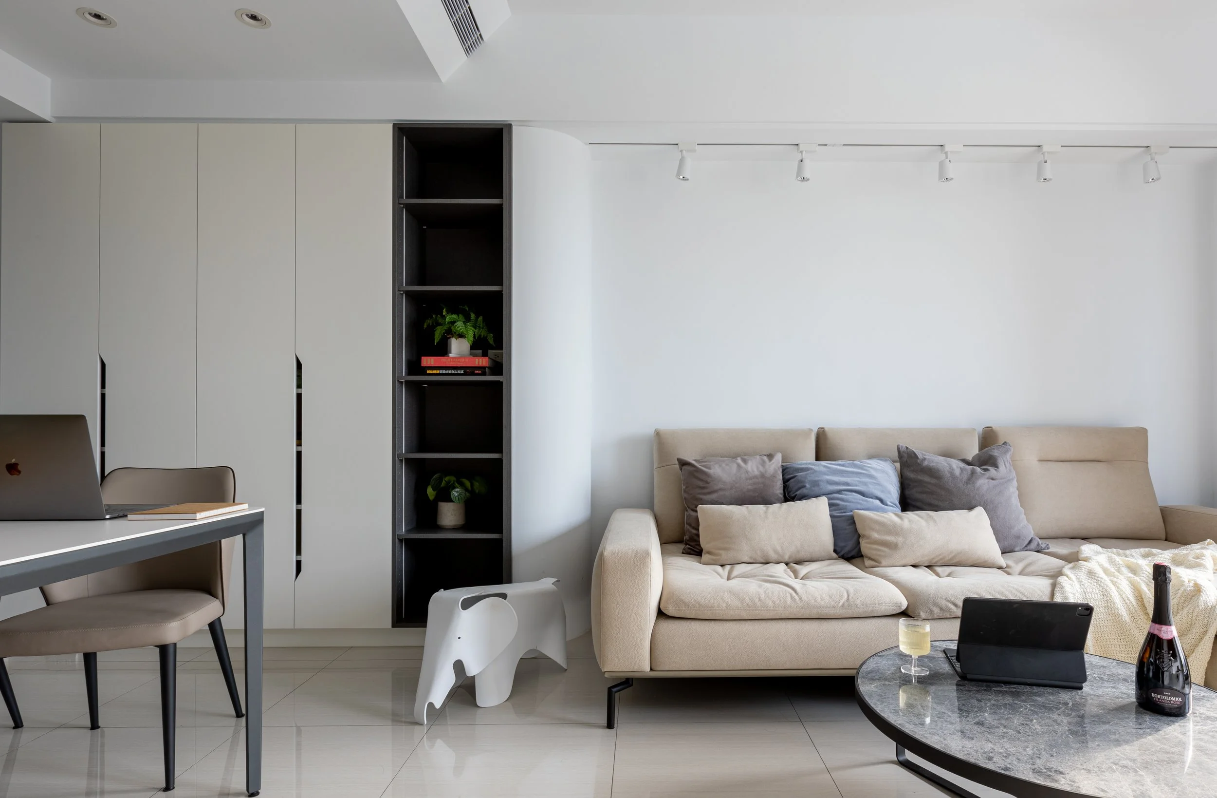Modern living room with beige sofa, multicolored cushions, black coffee table with a bottle, glass, and tablet, black shelving with plants and books, white built-in cabinets, and a small white elephant-shaped stool.