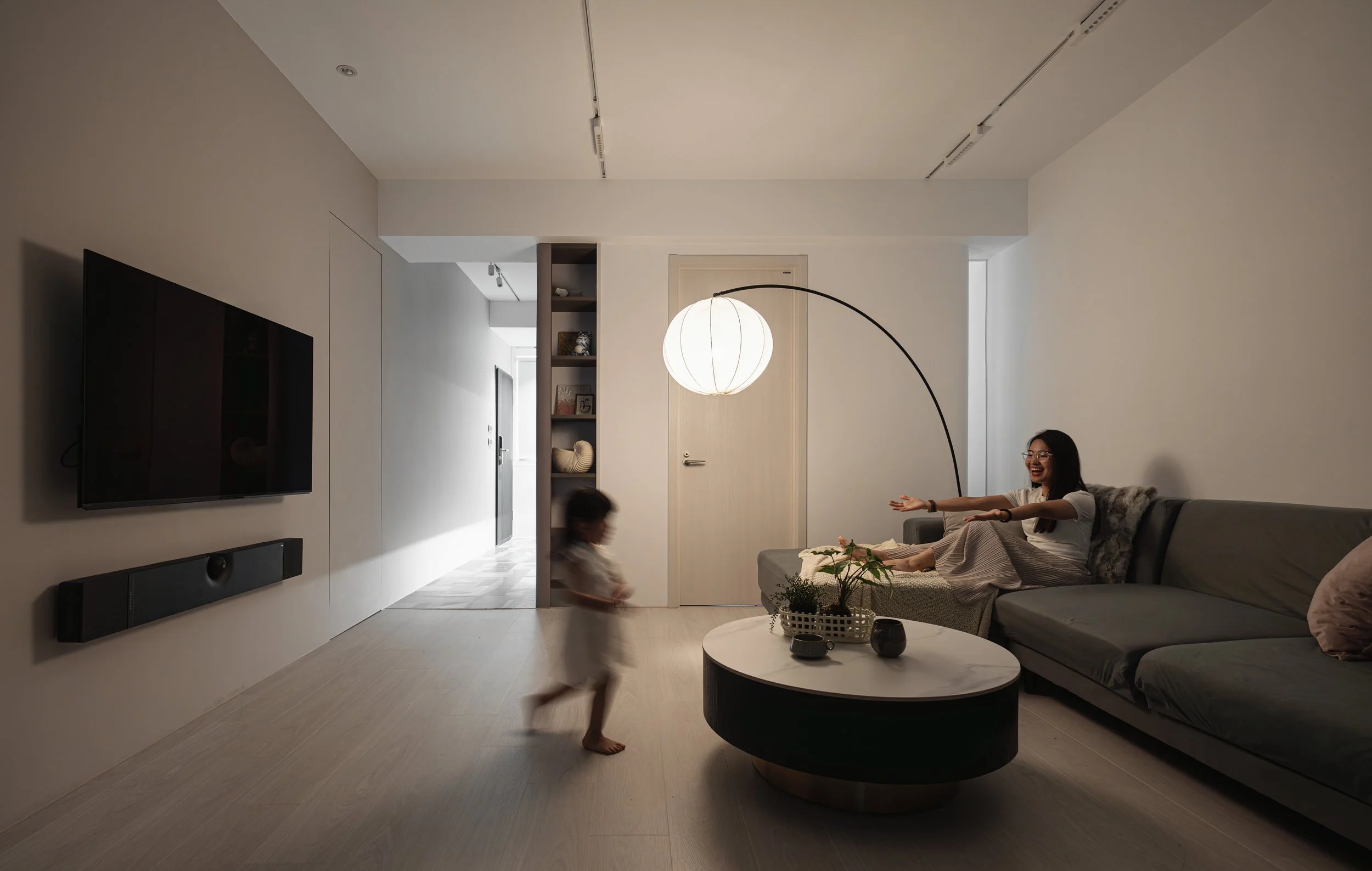Living room with a woman and child playing; woman sitting on a sofa stretching, child running, modern decor, wall-mounted TV, floor lamp, and minimal furniture.