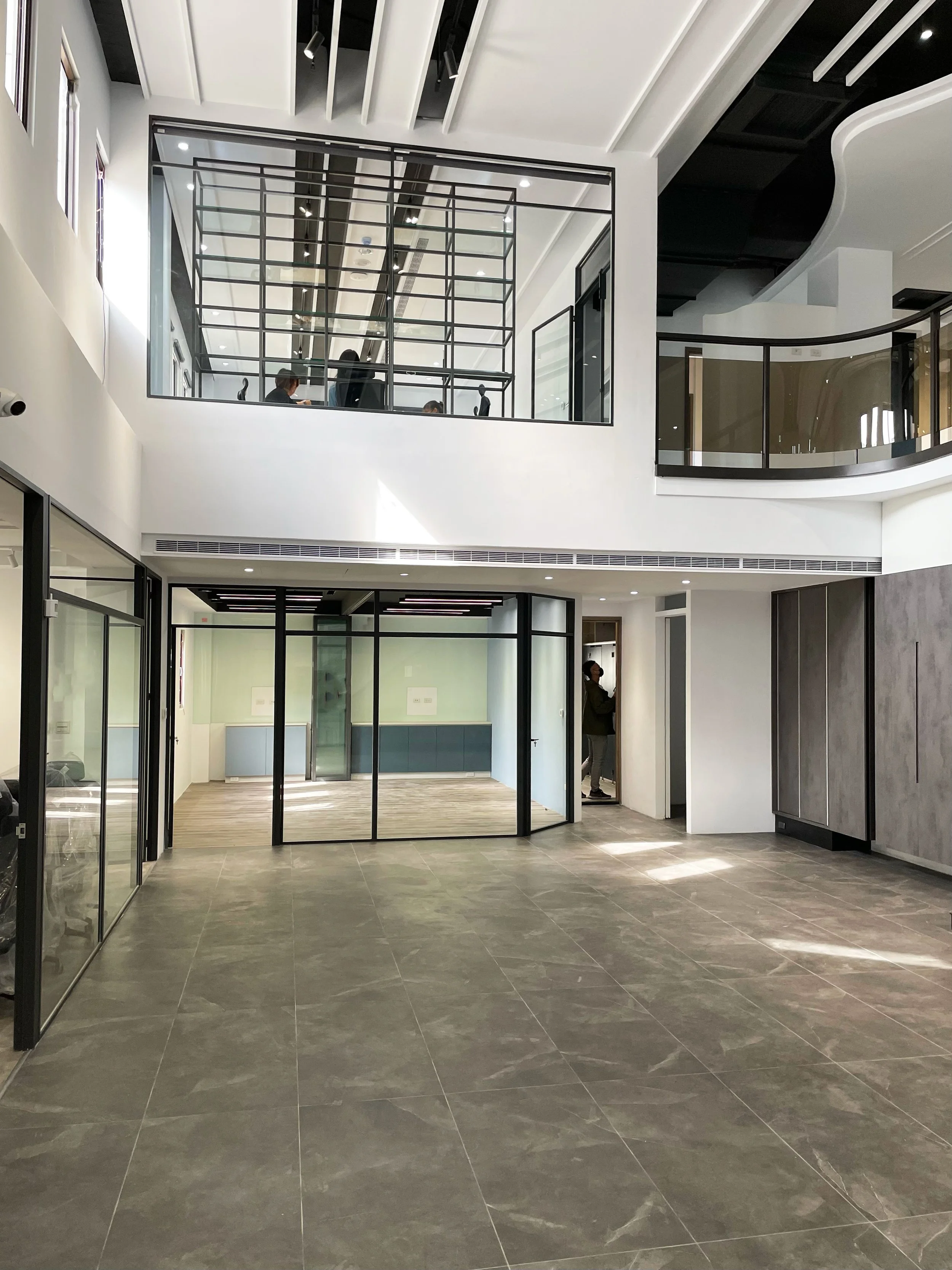 Modern office lobby featuring gray tiled floors, white walls, glass walls framed in black metal, and a double-height ceiling with white beams and an upper balcony area. The open and airy space welcomes visitors with sleek, contemporary design element