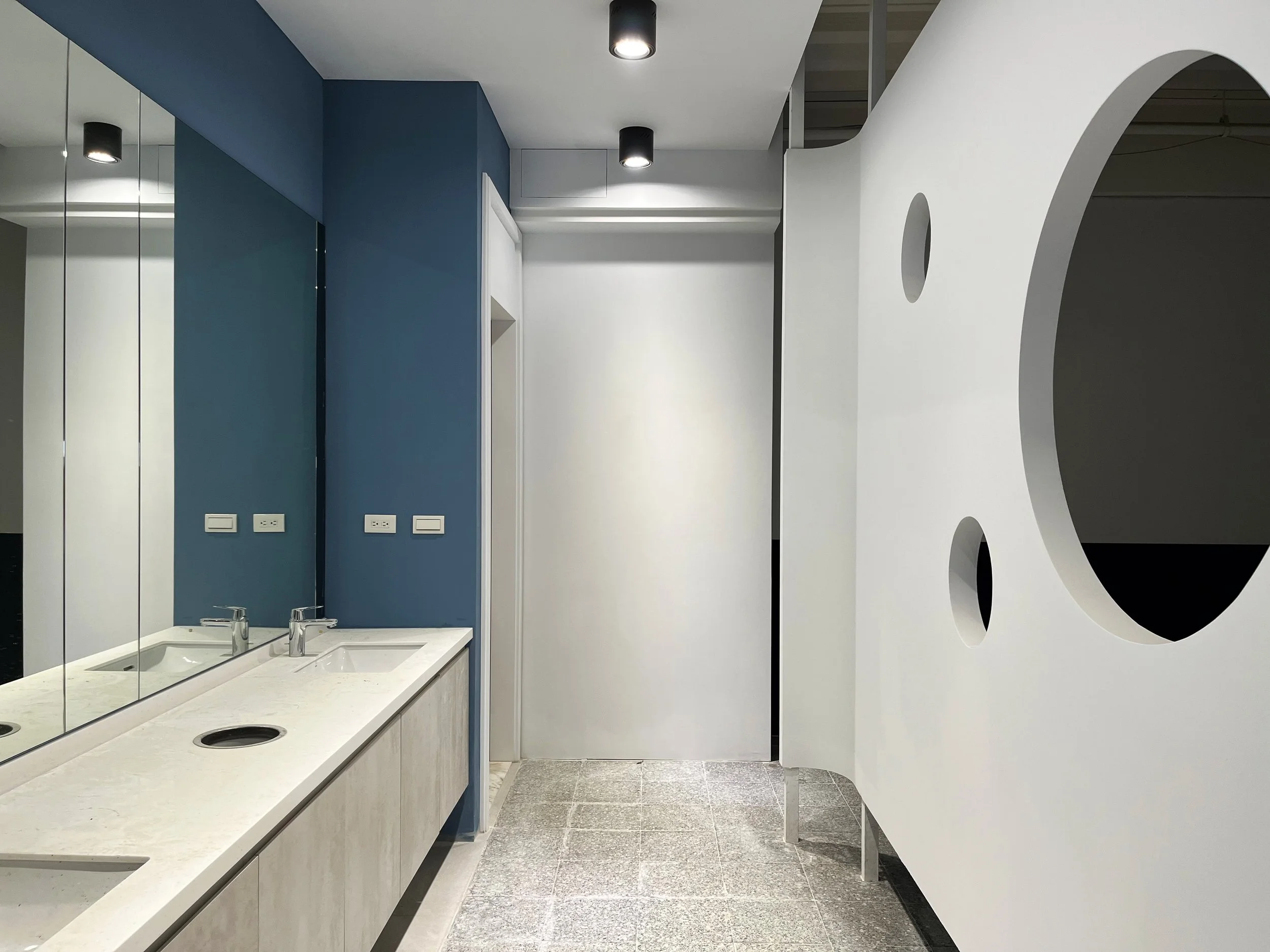 Modern public restroom with a double sink vanity and blue accent wall, mirror, and unique white partition with circular cutouts.