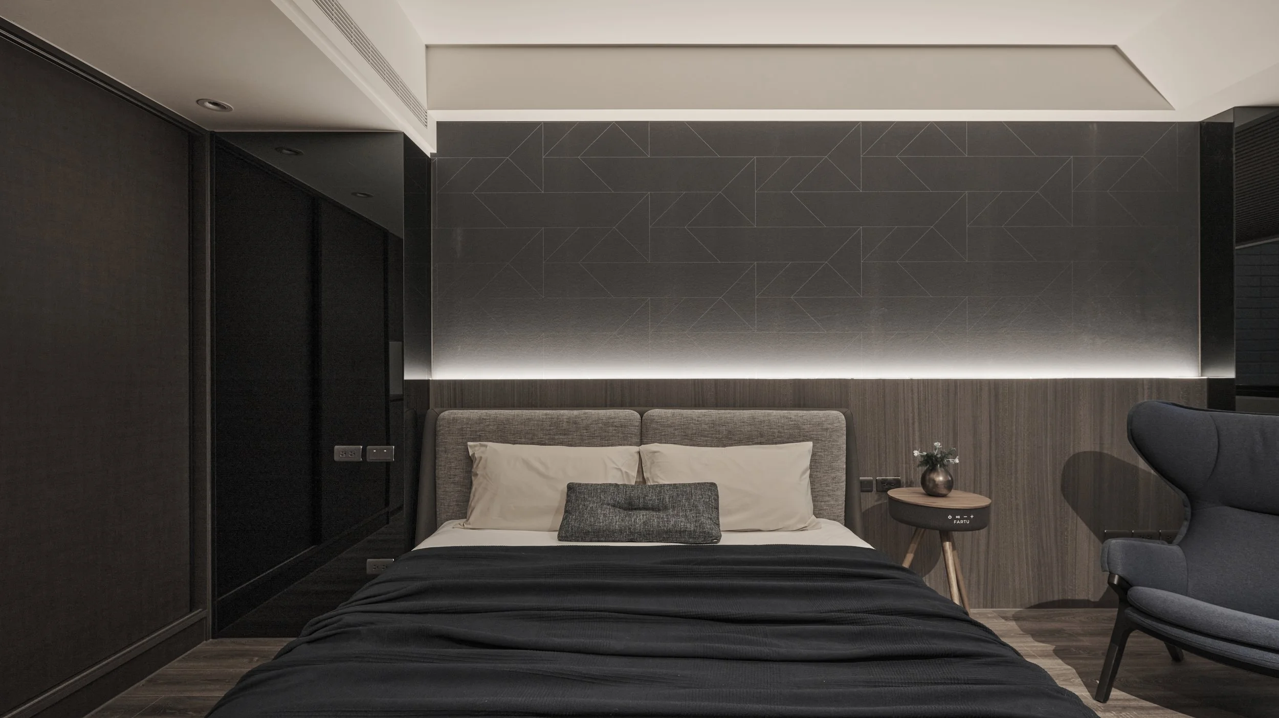 Modern townhouse bedroom featuring an upholstered bed with beige and gray decorative pillows, black bedspread, wooden nightstand with a small flower vase, black armchair, dark accent wall with subtle geometric pattern, and soft ambient lighting.