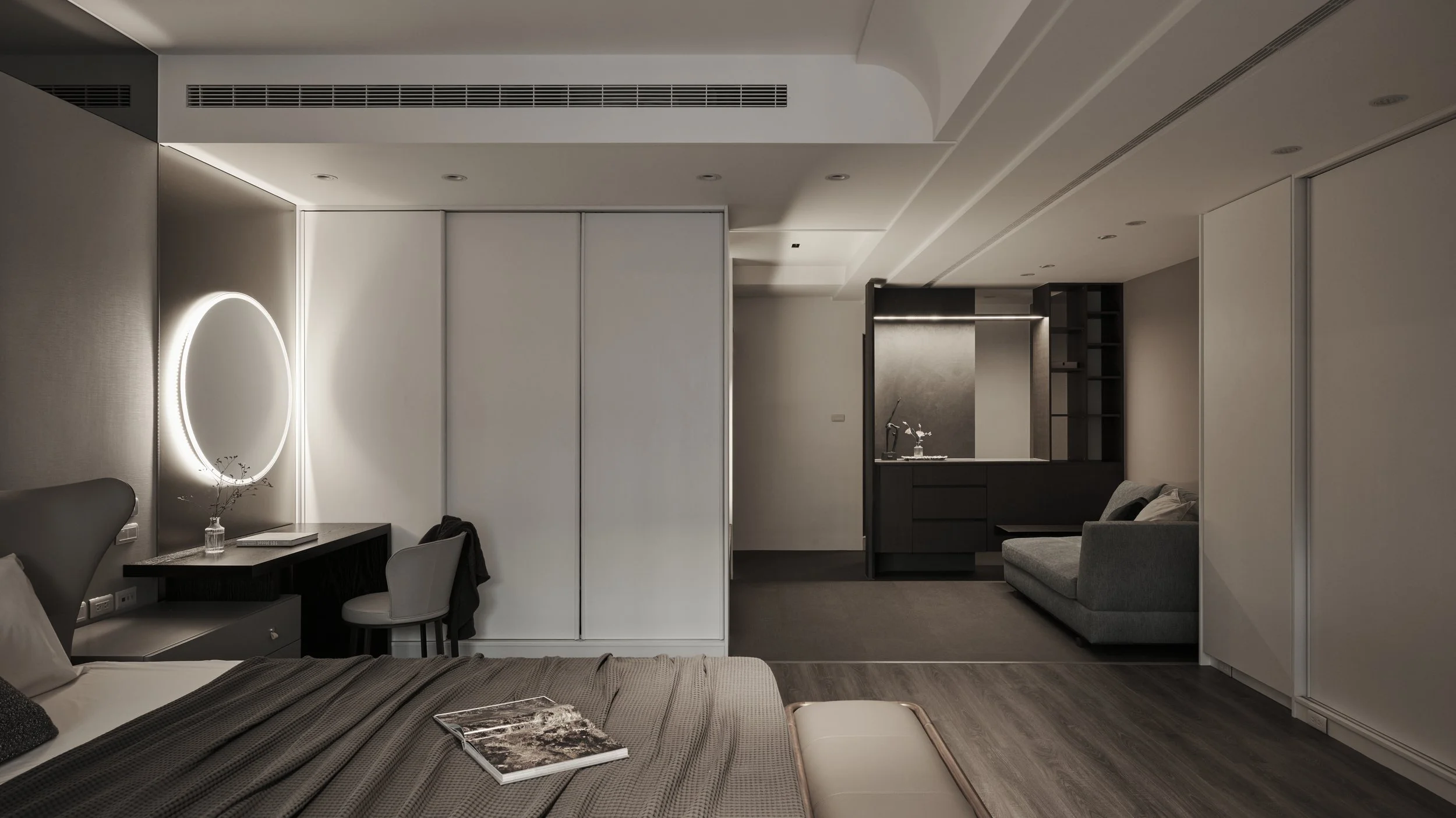 Modern hotel room interior with a bed, a illuminated round mirror, a small desk with a chair, and a sitting area with a sofa, in a minimalist design.