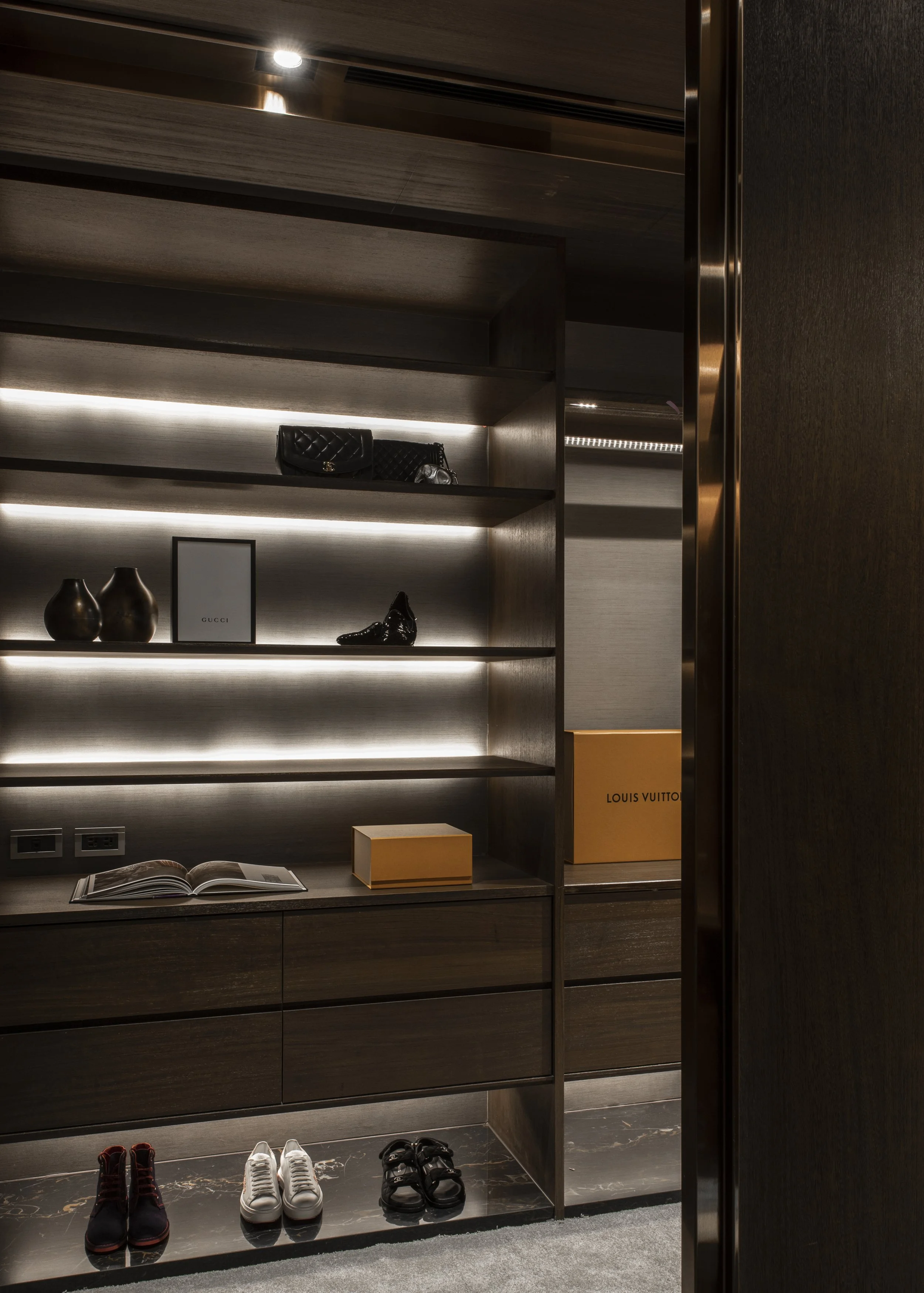Luxury walk-in closet featuring wooden shelves, designer shoes, handbags, decorative vases, and boxes, illuminated with LED lighting.