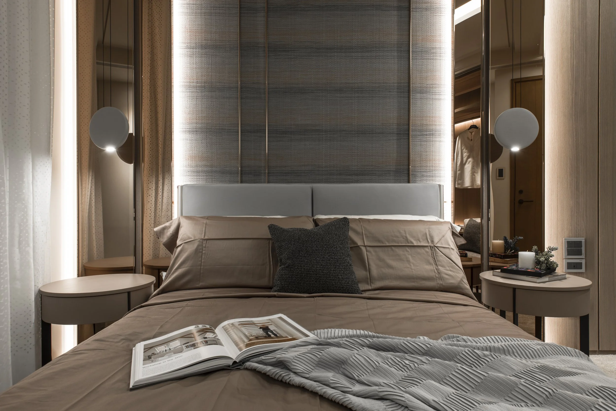 A modern bedroom with a neatly made bed, beige bedding, and a black throw pillow. On the bed, an open magazine is visible. The side tables have candles, books, and small decorative plants. There's a tall, ornate headboard behind the bed, flanked by a