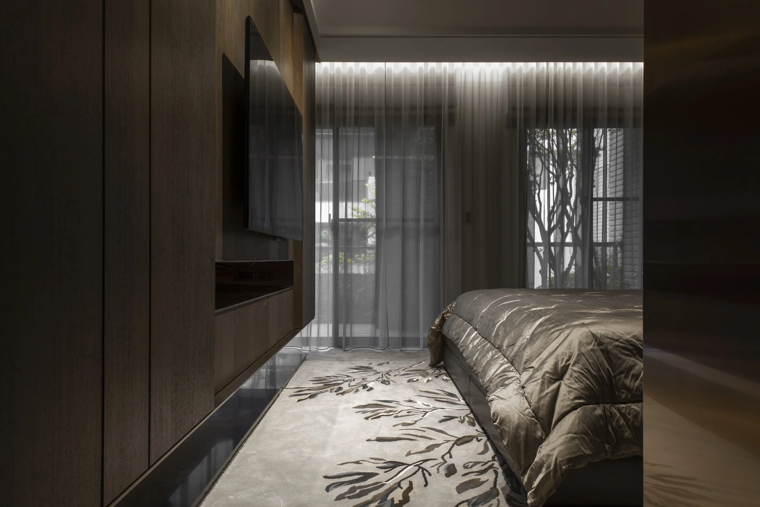 Modern bedroom with dark wood wall, flat-screen TV, beige patterned rug, and a bed with a shiny brown comforter, large windows with sheer curtains, and a tree visible outside.
