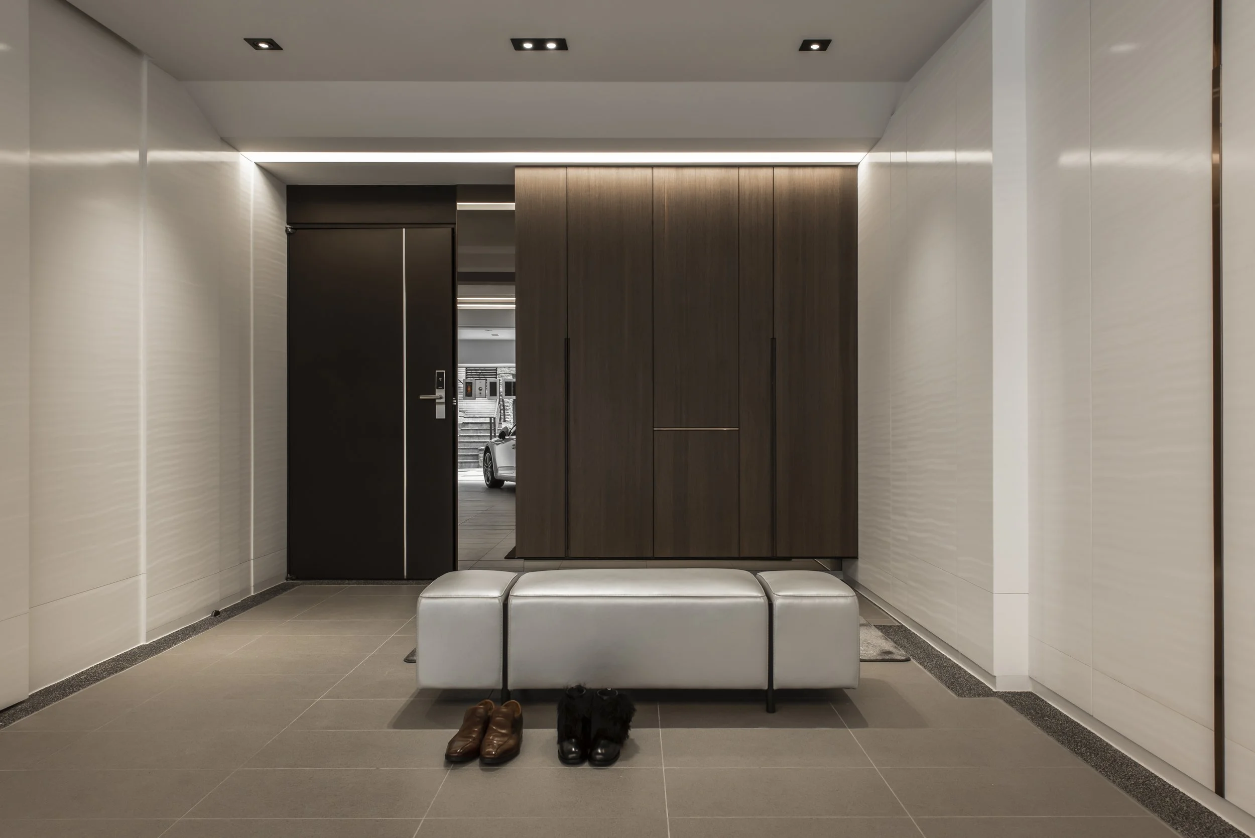 Modern garage lobby featuring a white bench with neatly placed brown and black shoes underneath, wooden and white walls, and a door leading outside where a car is visible.