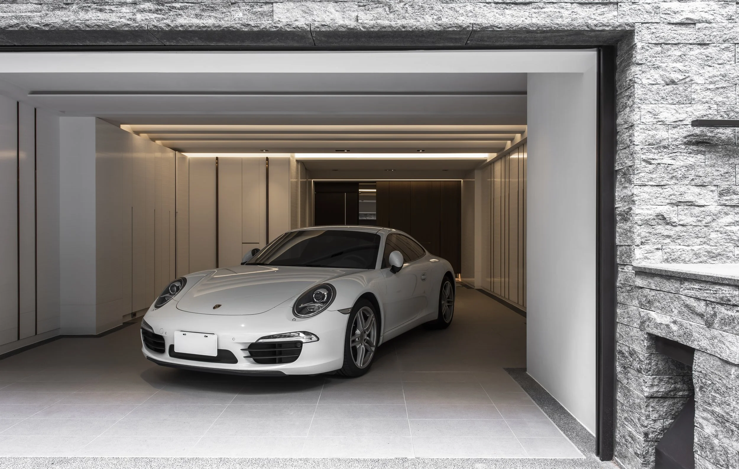 A white sports car parked inside a modern garage with stone and white wall accents and ceiling lighting.