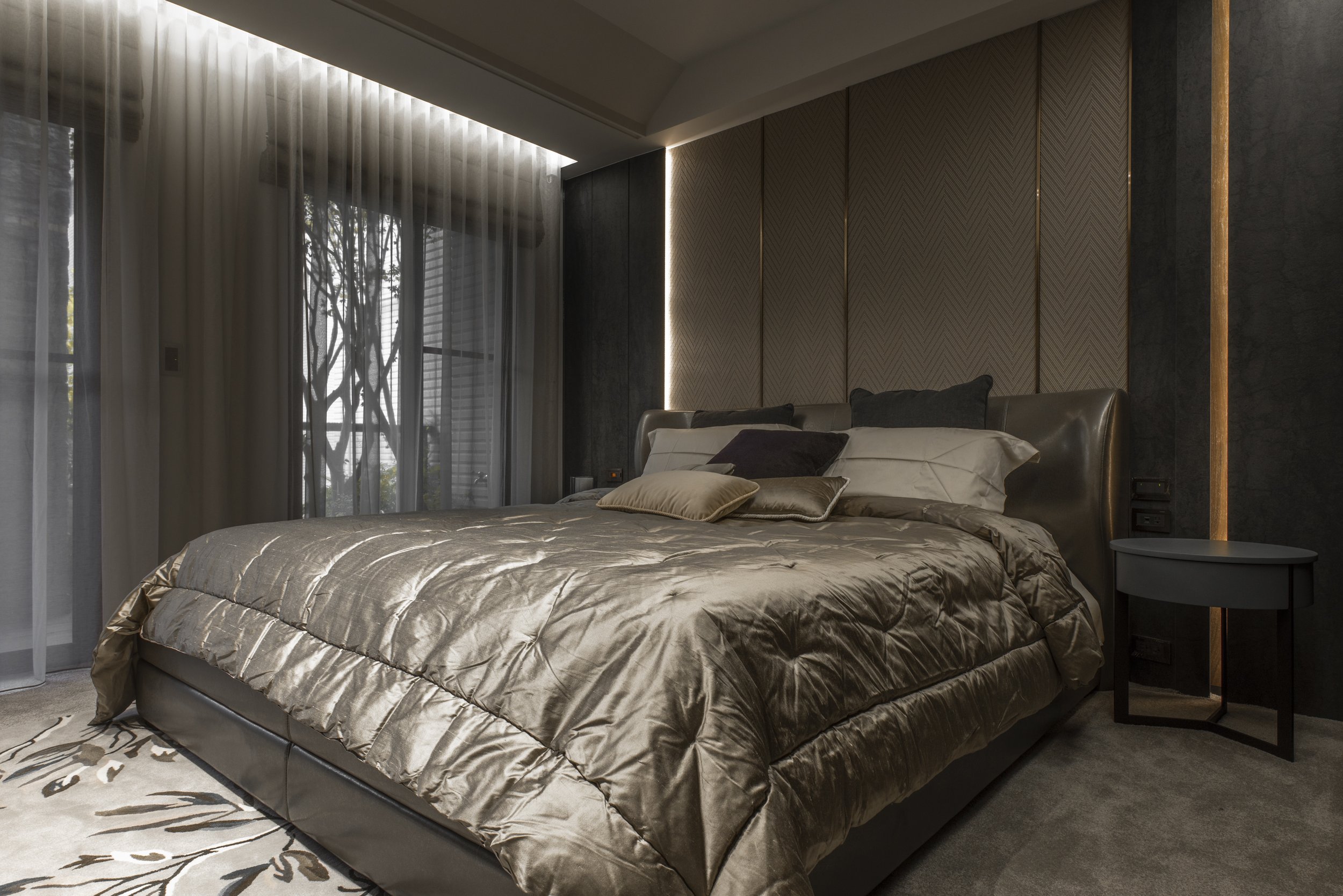 Luxurious bedroom with a large, satin gold bed, multiple pillows, a dark side table, and floor-to-ceiling sheer curtains, with natural light filtering through and modern decor elements.