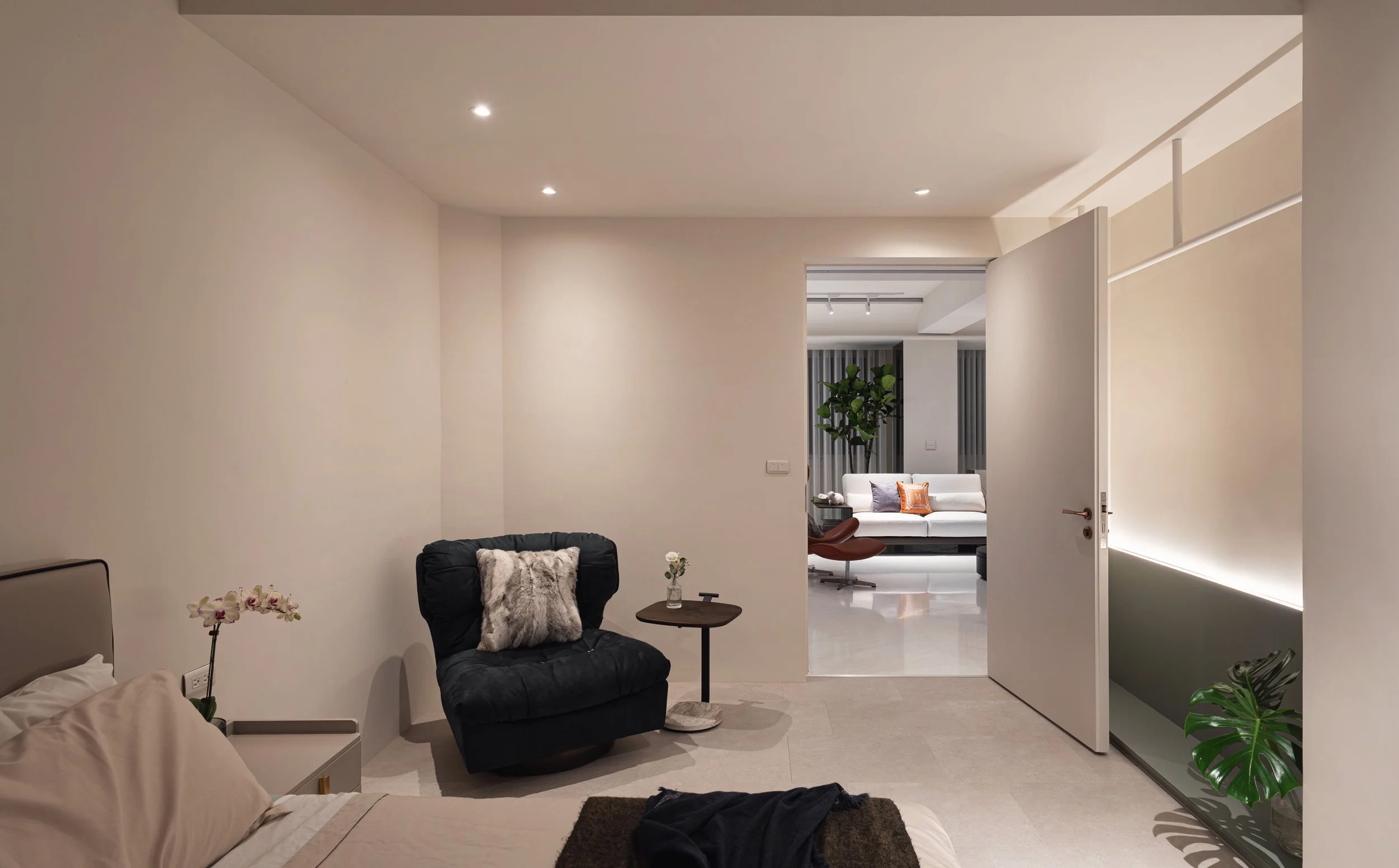 Modern bedroom with beige bed and pillow, black armchair with fur pillow, small side table with a vase of flowers, open door revealing a white sofa with cushions, green plant, and contemporary decor.