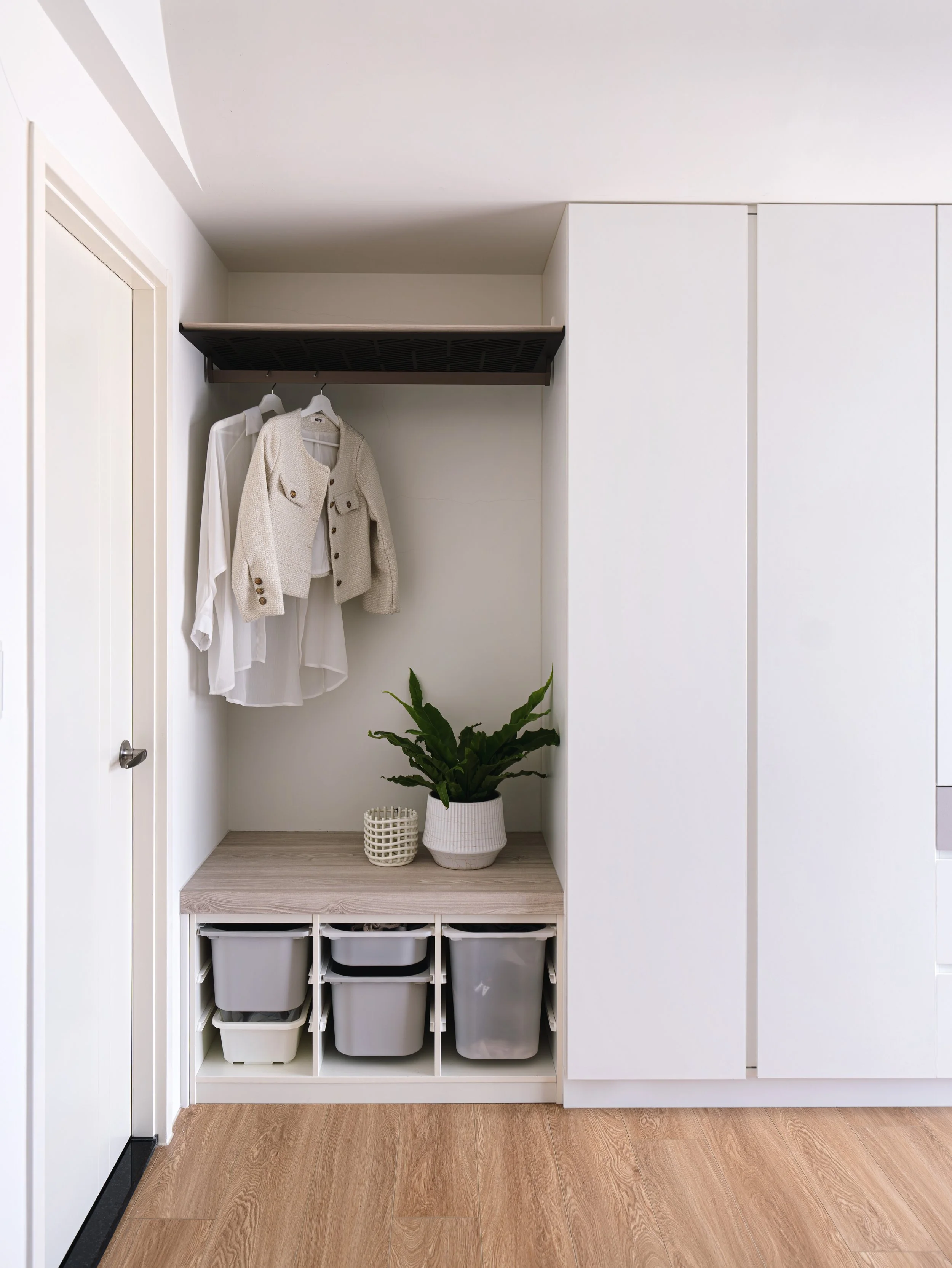 Modern minimalist entryway with hanging white and beige coats on a black shelf, a large green plant in a white pot, and storage bins underneath on a light wooden floor.