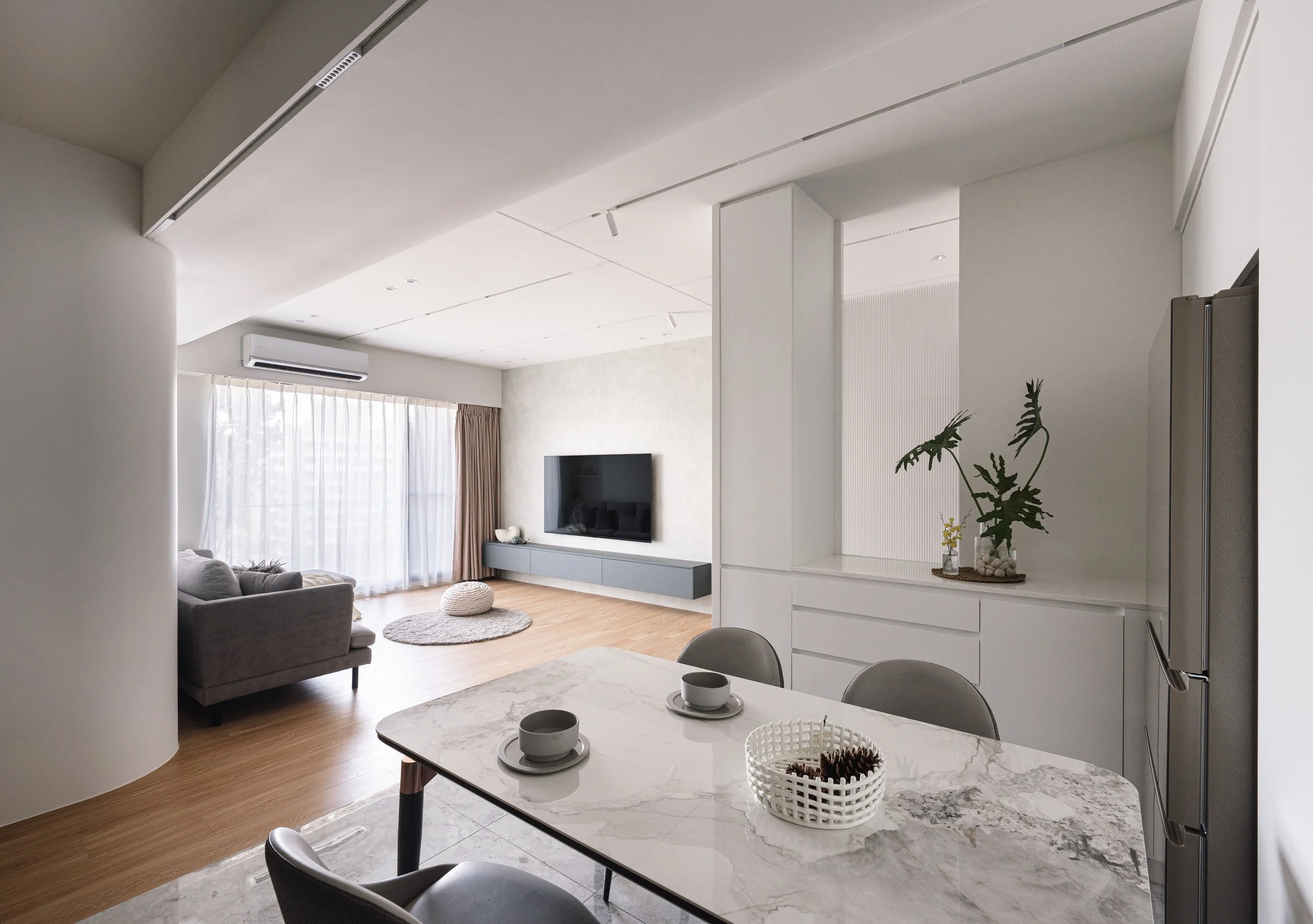 Modern living room with a sofa, TV, and a window with curtains, seen from the dining area with a marble table.