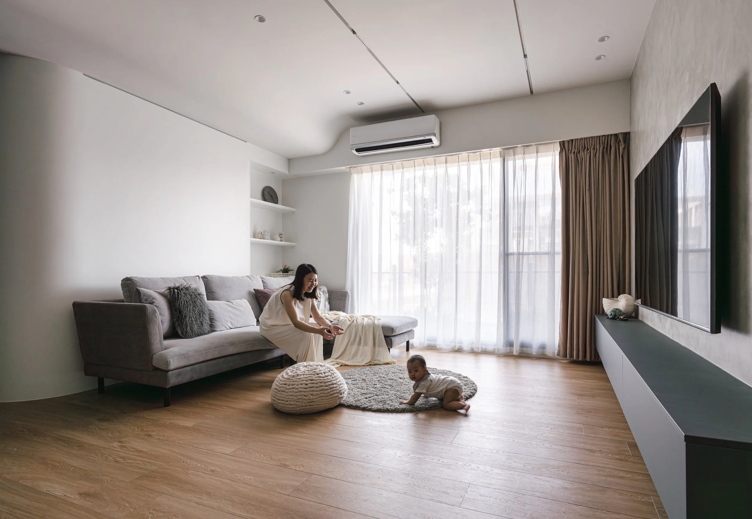 A woman and a baby playing on a wooden floor in a living room with a large gray sofa, a knitted pouf, a small round rug, and a wall-mounted TV.