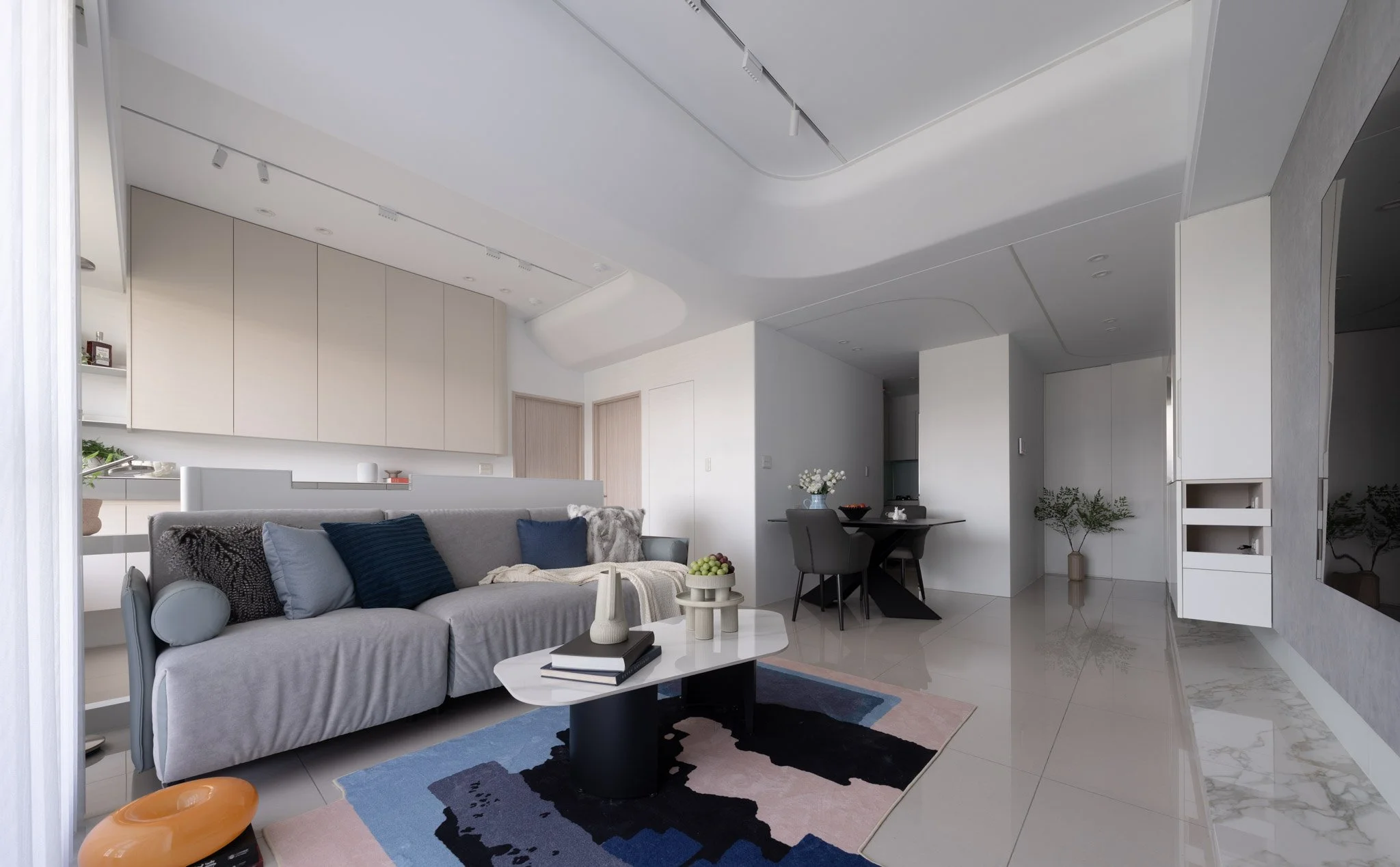 Modern living room with a grey sofa, colorful pillows, a white coffee table with decorative items, a dark dining table with chairs, potted plants, and a large flat-screen TV, all in a minimalist design with light-colored walls and flooring.