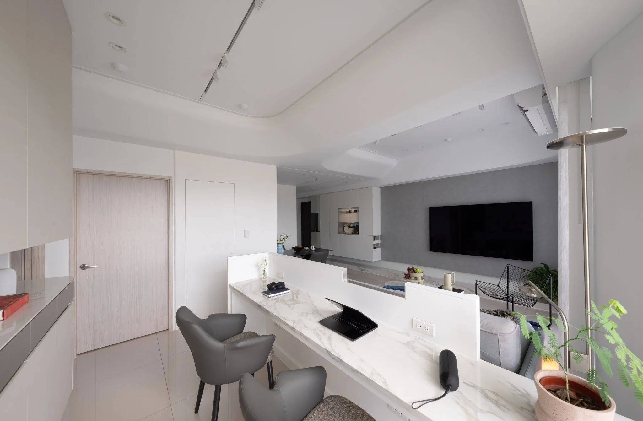 Modern living room with white walls, a mounted flat-screen TV, gray chairs, a marble countertop with a laptop, potted plants, and a seating area.