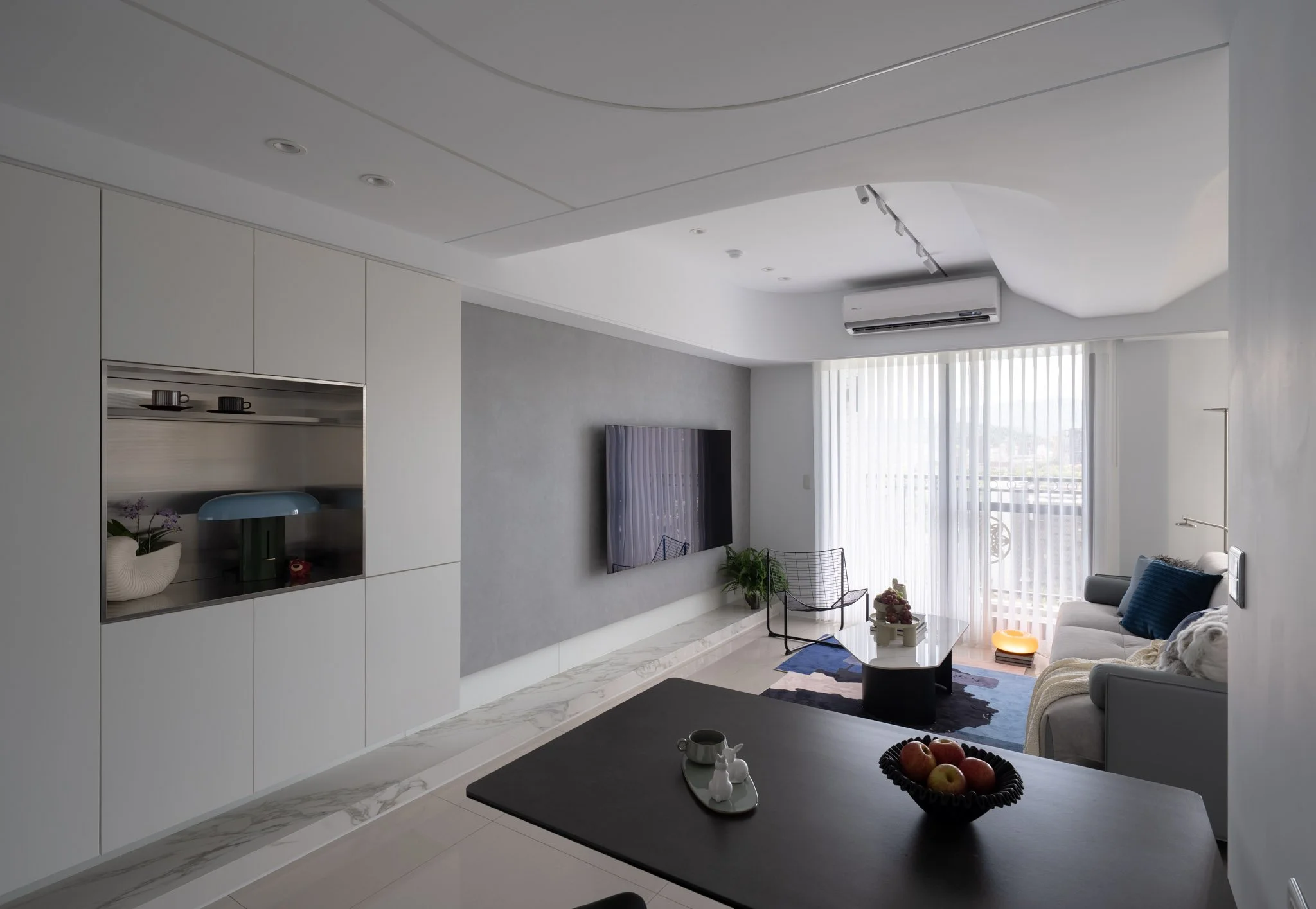 Modern living room with a white sofa, black wire chair, flat-screen TV on gray wall, and large window with vertical blinds.