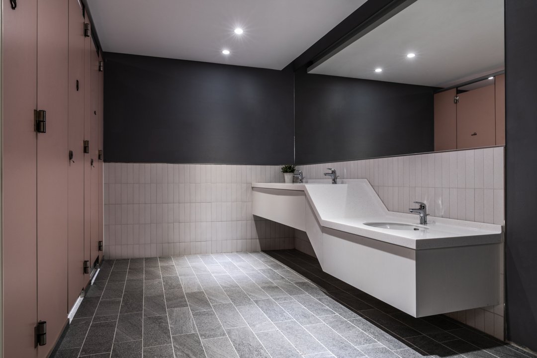 Public restroom with pink lockers, dark gray walls, white tiled wainscoting, gray tiled floor, and two sinks with faucets under a large mirror.