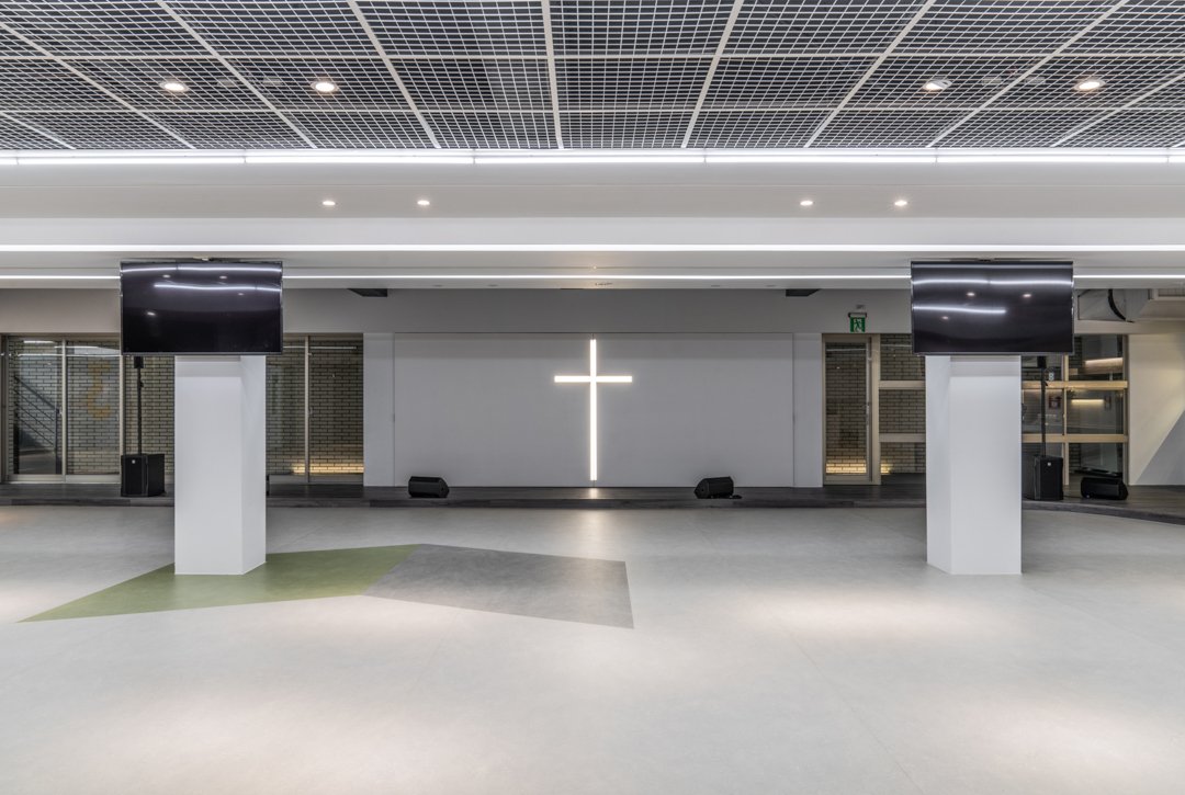 Modern church interior with illuminated cross on the back wall, two large screens on columns, and microphones on the stage.