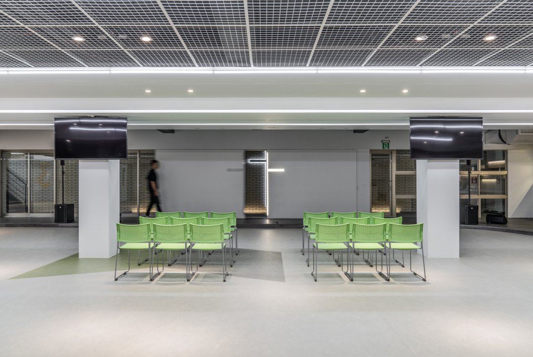 Chapel interior featuring rows of green chairs, an illuminated cross on a brick wall, two large black screens, and mounted speakers, creating a serene and contemplative atmosphere.