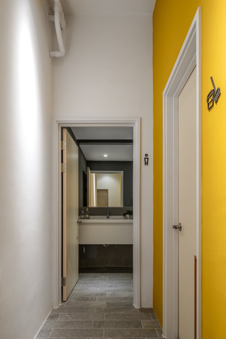 A hallway leading to a bathroom, with a yellow wall on the right, a white wall on the left, and a mirror reflecting the hallway. The bathroom has a white sink, dark tiles, and a plant on the counter.