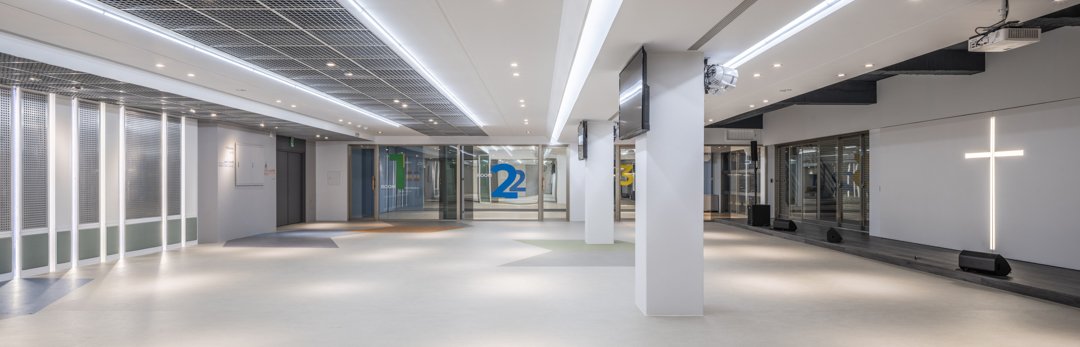 Modern church lobby featuring white walls, ceiling lighting, expansive glass walls and doors, a prominent cross on the wall, and numbered signs—blending spiritual significance with contemporary architectural design.