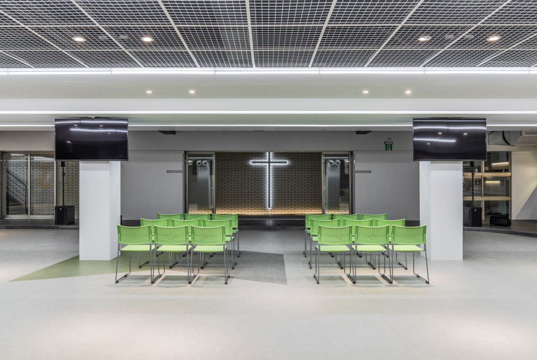 Chapel interior featuring rows of green chairs, an illuminated cross on a brick wall, two large black screens, and mounted speakers, creating a serene and contemplative atmosphere.