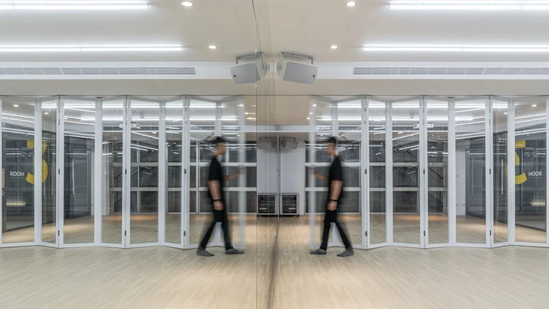 A modern church multipurpose room, featuring wood flooring, glass walls, ceiling lights, and a full-length rehearsal mirror. A person walks past, highlighting the space's dynamic use for activities such as dance practice and group events.