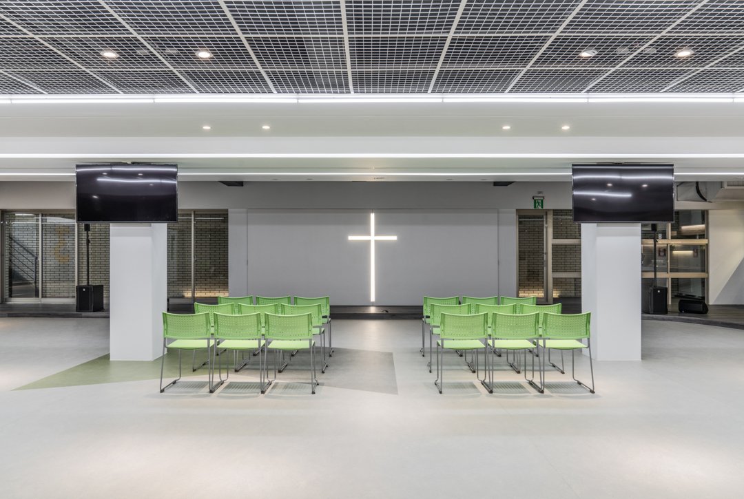 Chapel interior featuring rows of green chairs, an illuminated cross on a brick wall, two large black screens, and mounted speakers, creating a serene and contemplative atmosphere.