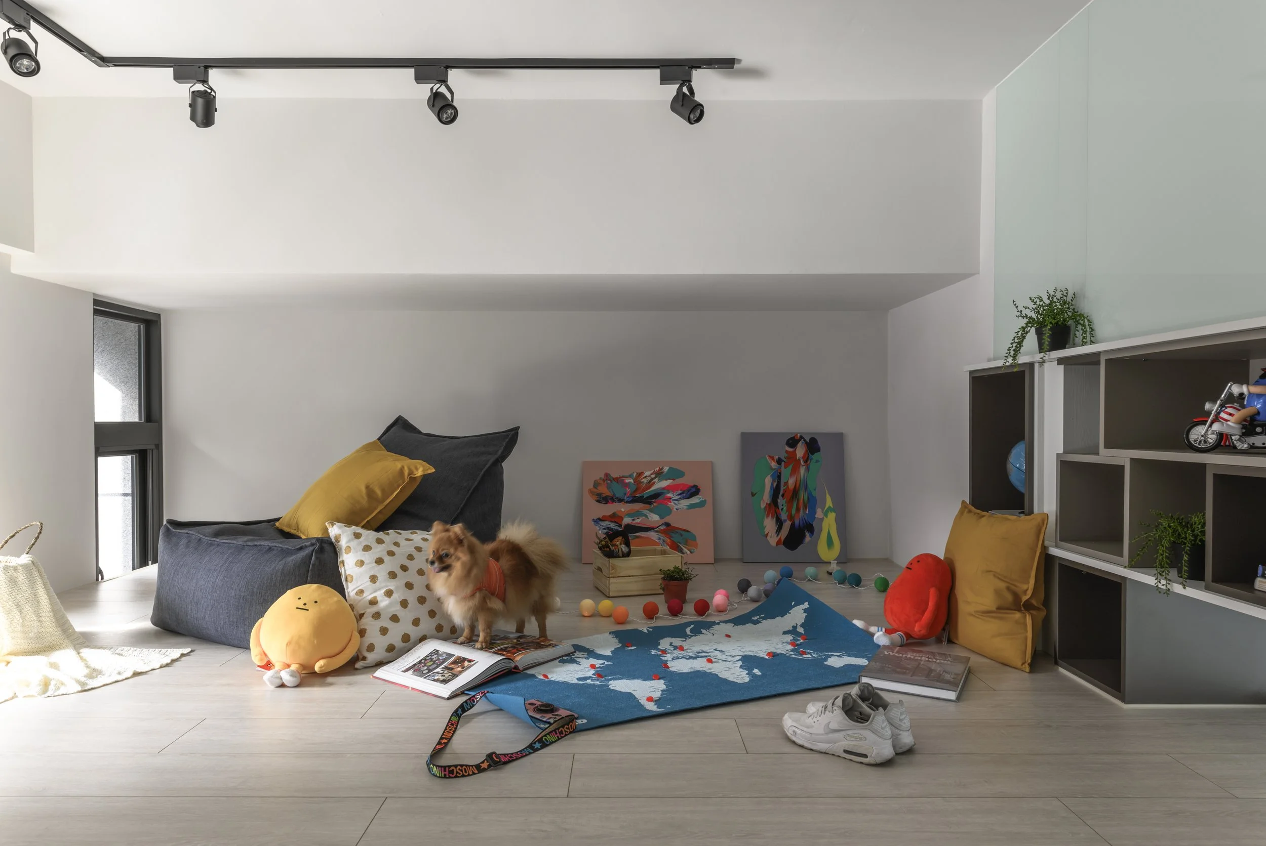 A cozy, modern living room corner with scattered toys, colorful artwork, a small dog, and cushions, illuminated by natural light from a window.