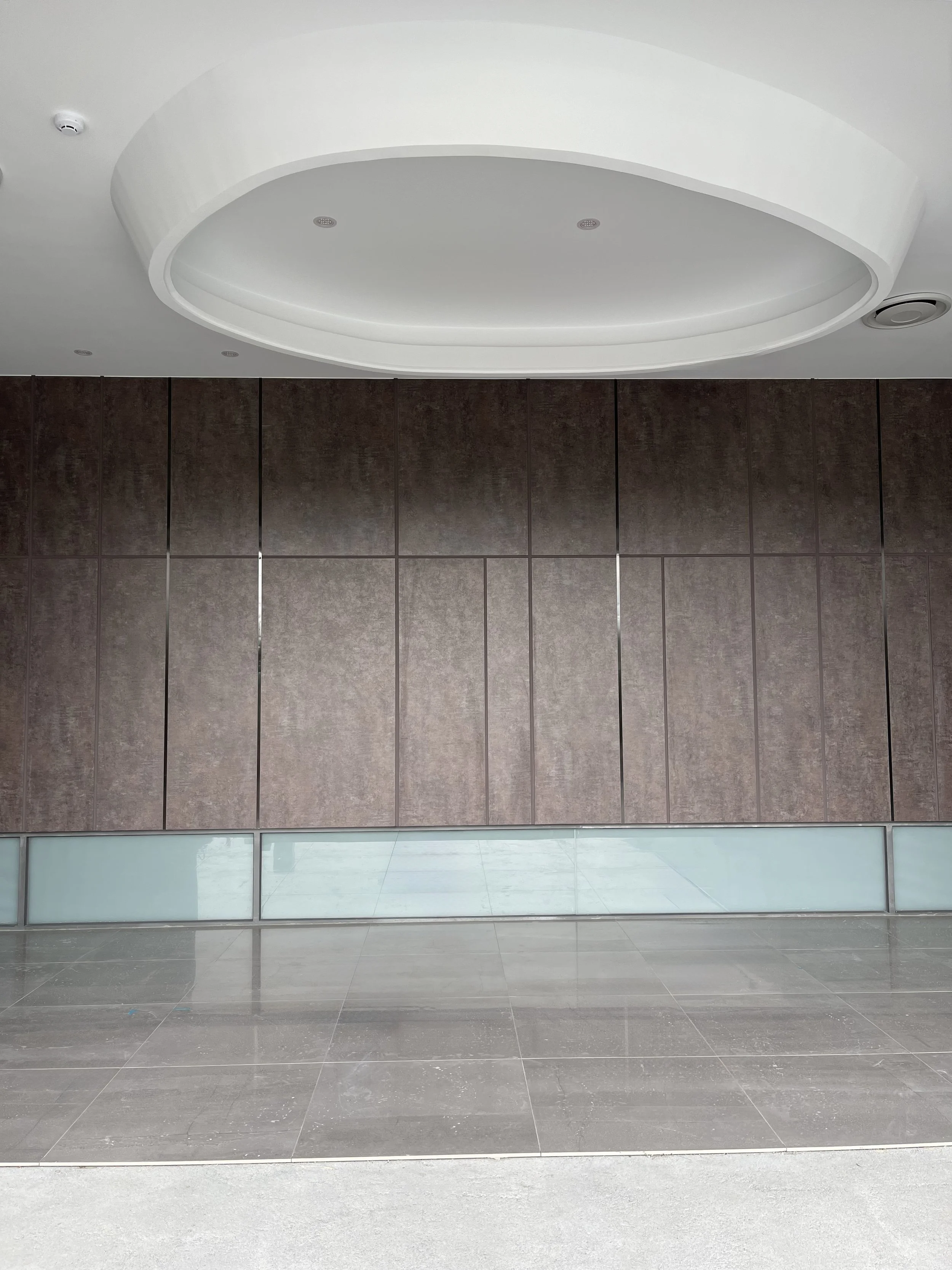 Interior of a modern building with large brown wall panels, a reflective tiled floor, a glass panel near the wall, and a white oval ceiling feature with small recessed lights.