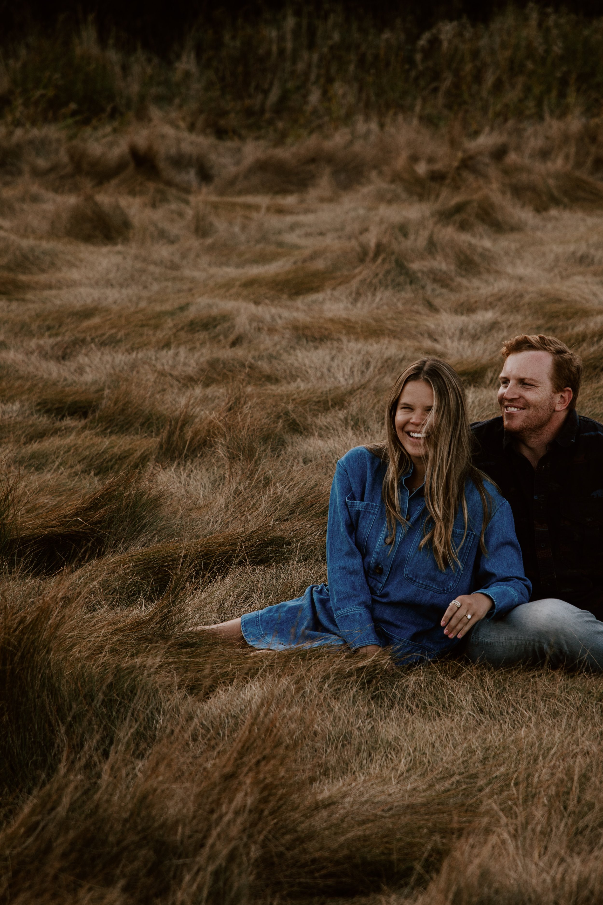A smiling couple sitting on a grassy field, with the woman wearing a blue denim shirt and the man in a dark shirt, enjoying each other's company.