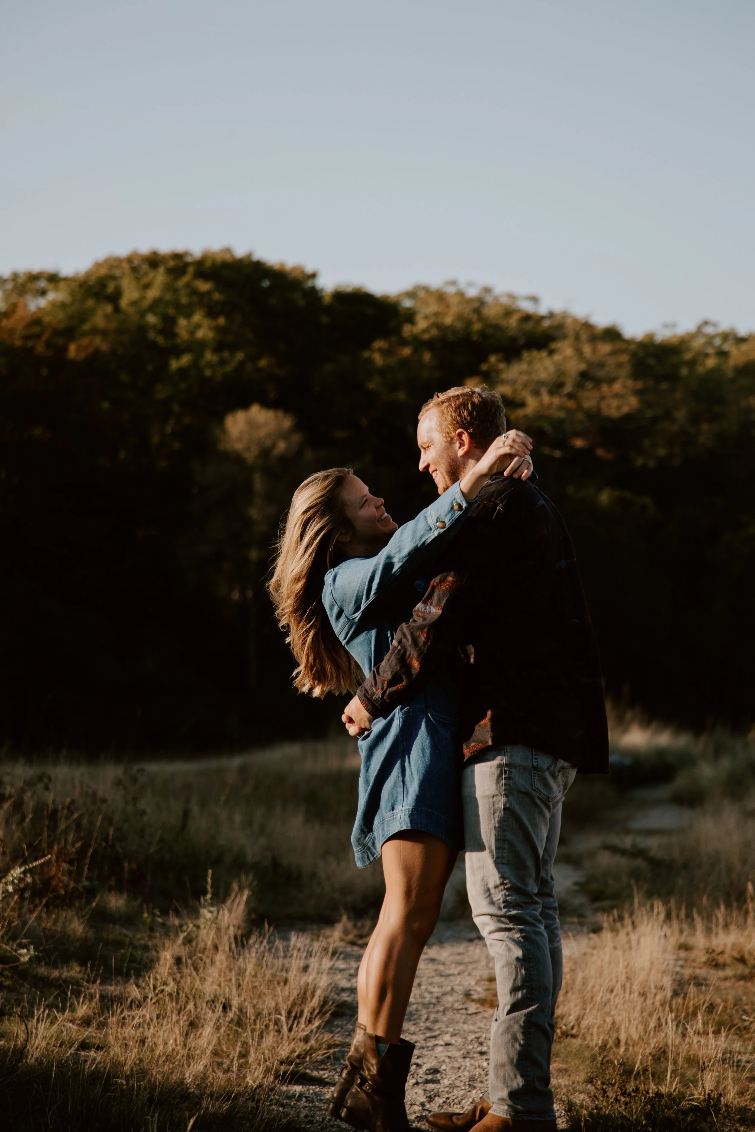 A couple is embracing in an outdoor setting with trees in the background during sunset.