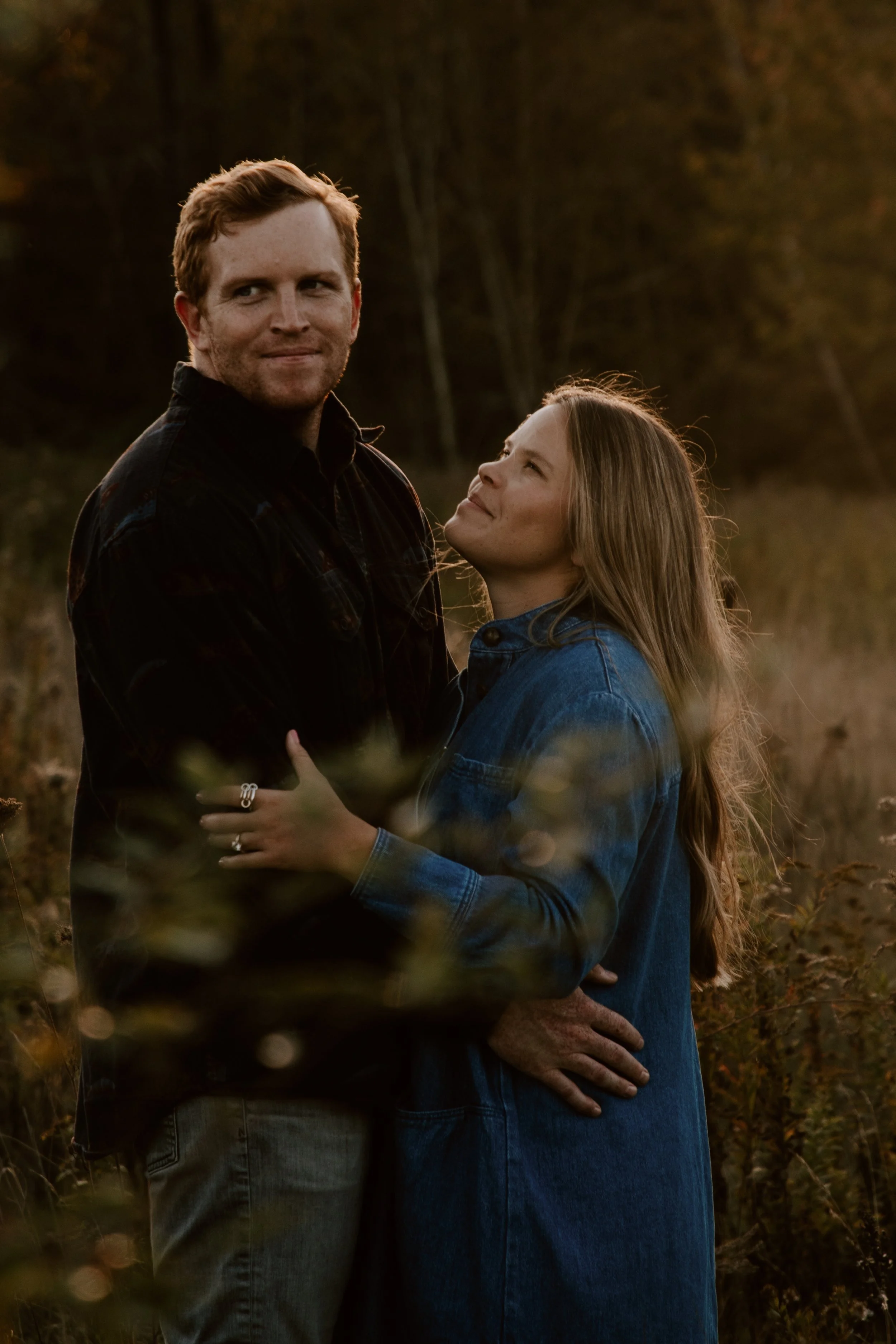 A couple standing outdoors in a field during sunset, embracing and looking at each other, with trees in the background.