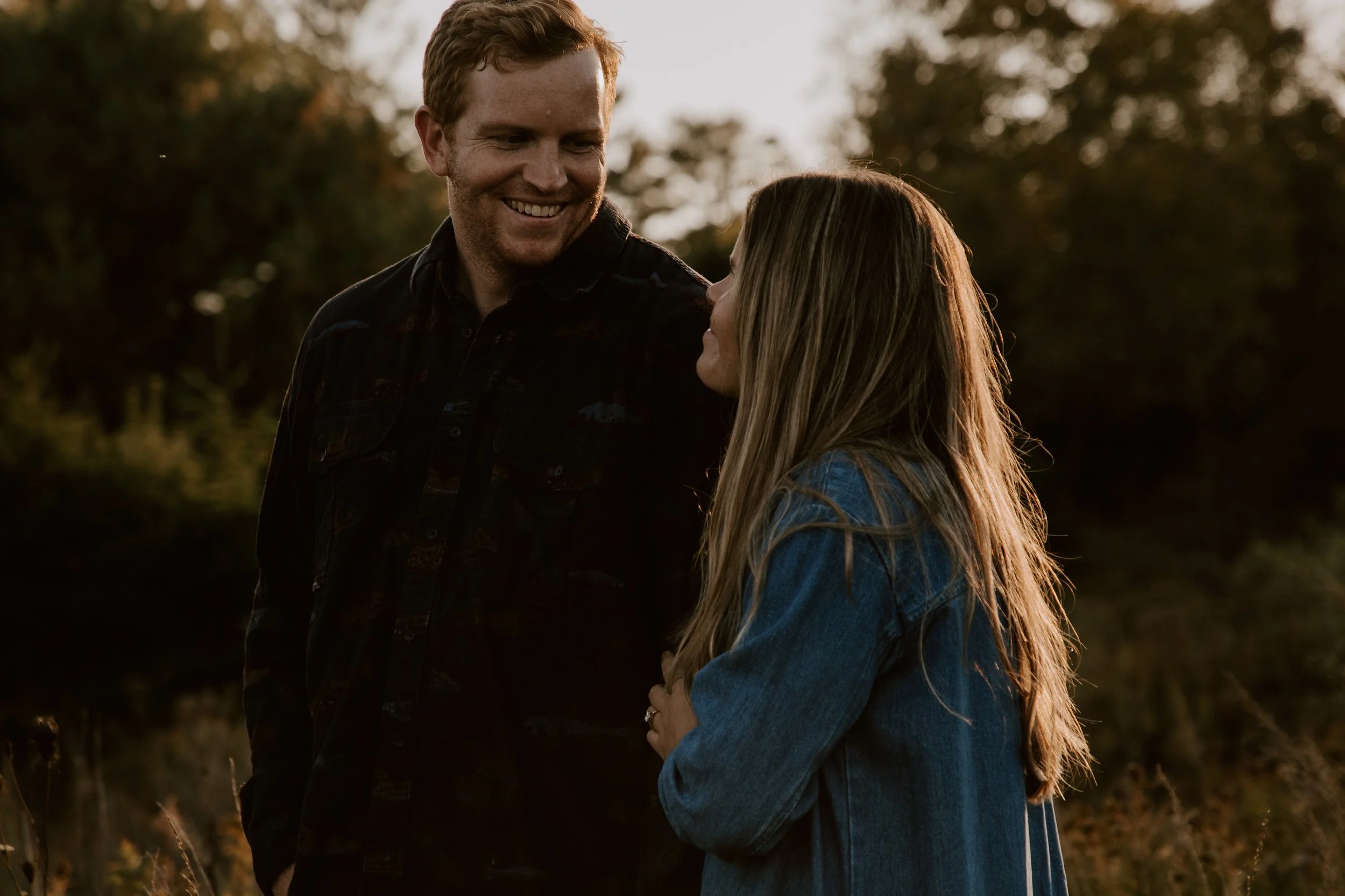 A man and woman smiling at each other outdoors during sunset, with trees and natural scenery in the background.