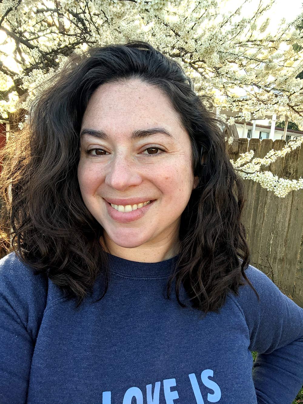 A woman with dark wavy hair smiling in front of blossoming white flowers on a tree, with a wooden fence and a house in the background.