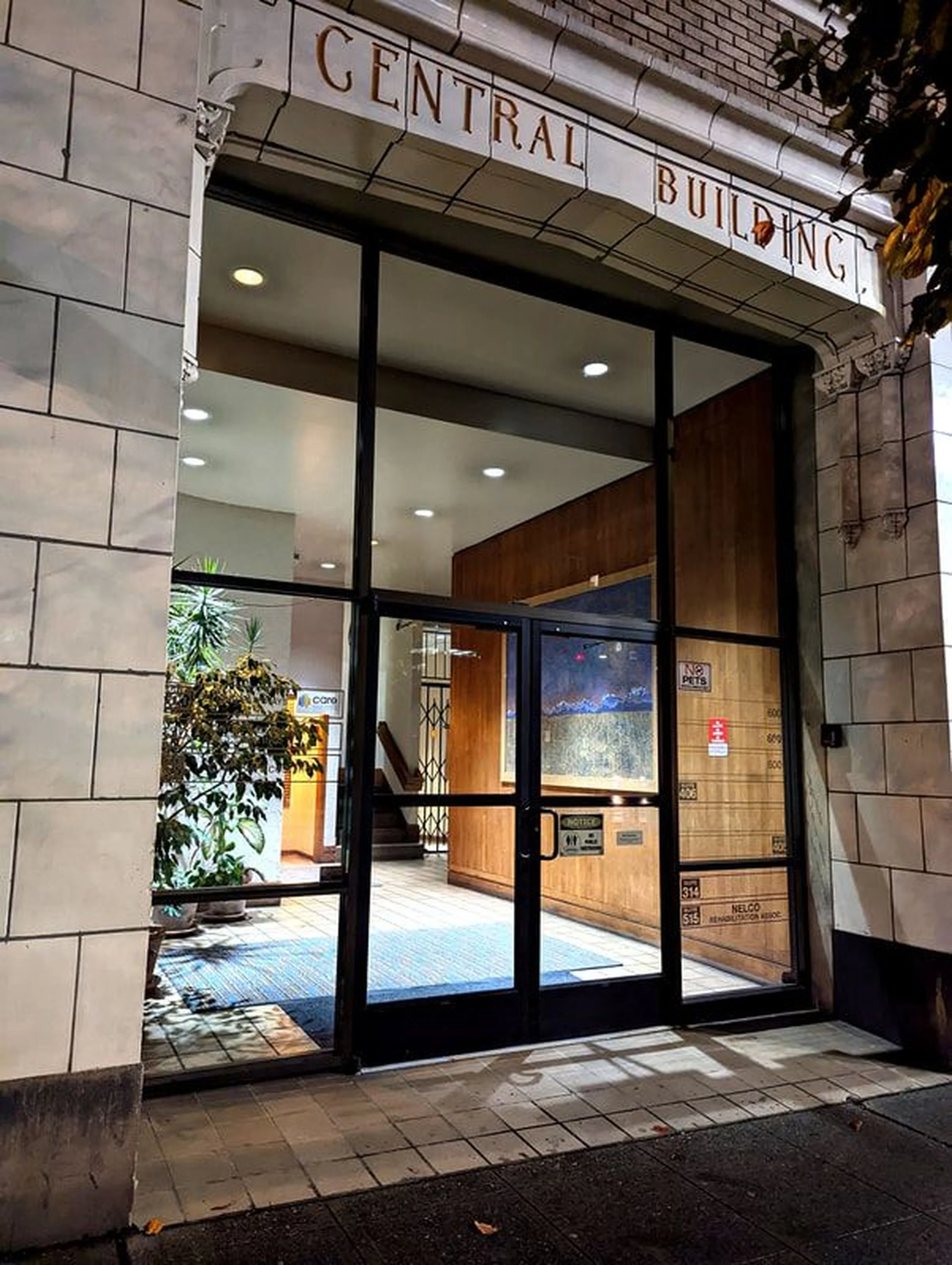 Night view of the entrance to a building labeled 'Central Building' with glass doors and windows, potted plants on the inside, and a staircase visible within.