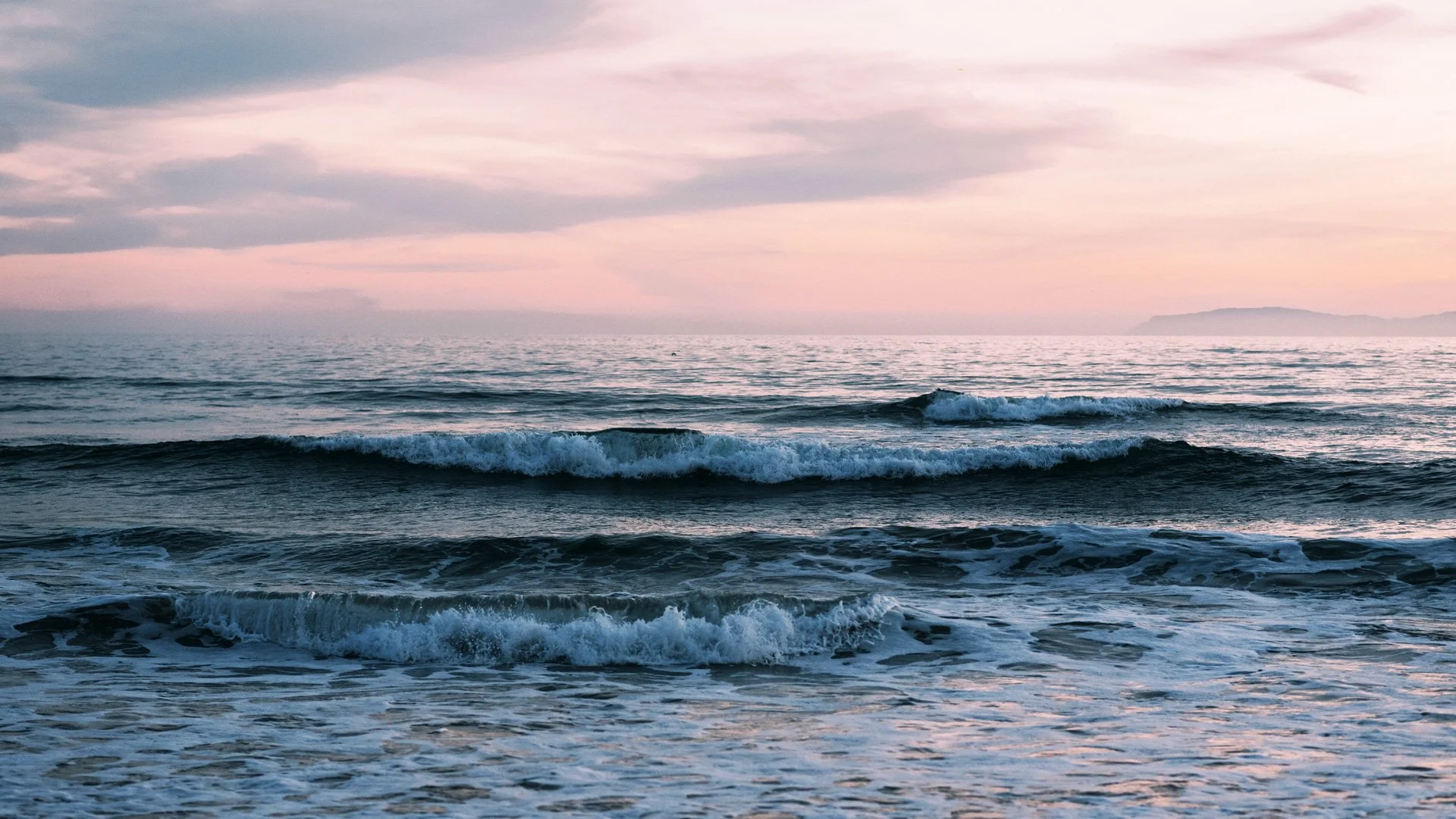 Sunset over the ocean with gentle waves crashing on the shore, pastel sky with pink and purple clouds, and distant landmass on the horizon.