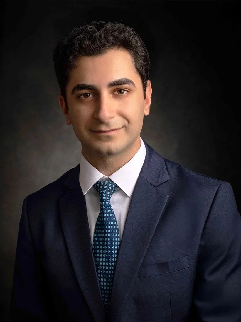 A young man with dark, curly hair in a navy blue suit, white shirt, and patterned tie posing against a dark background.