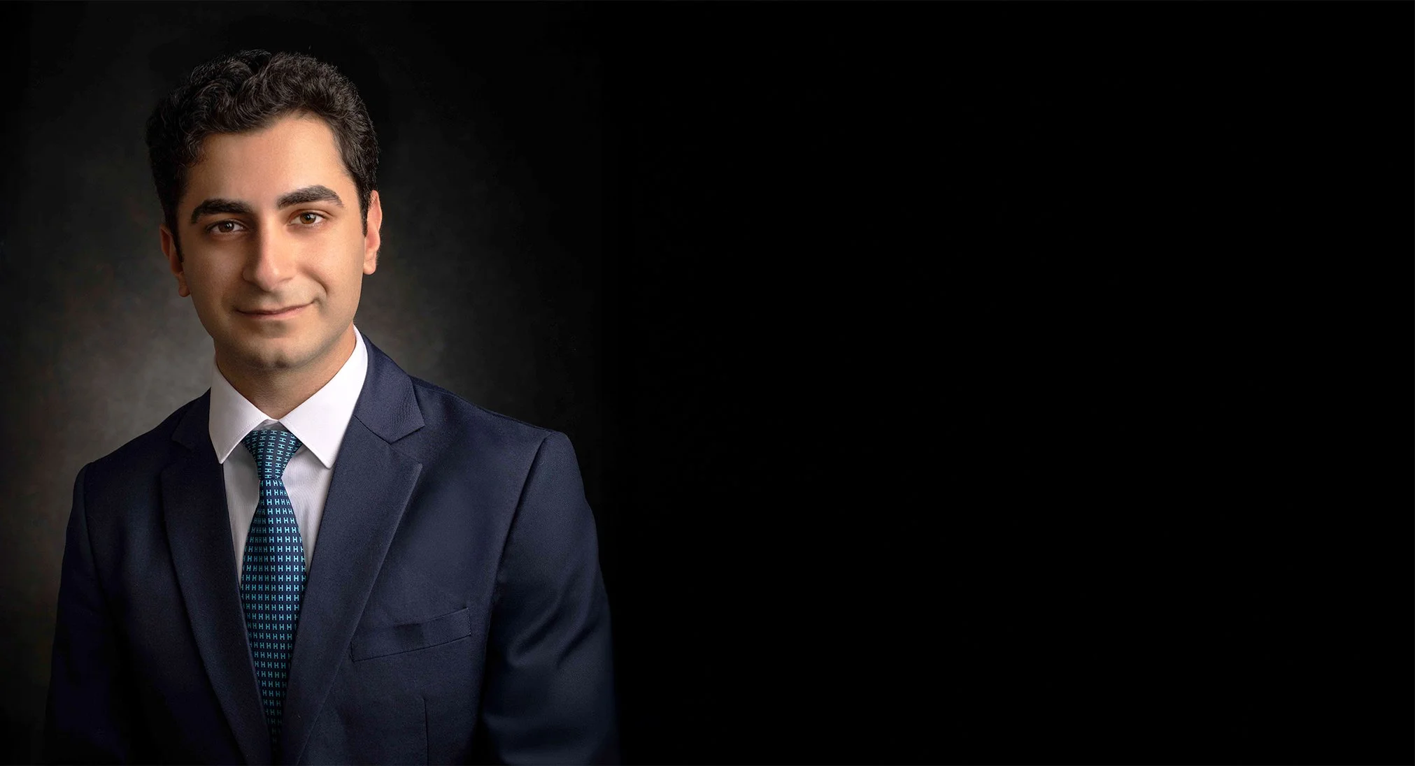 A young man with dark hair, dressed in a navy blue suit with a white shirt and patterned tie, is smiling slightly against a dark background.