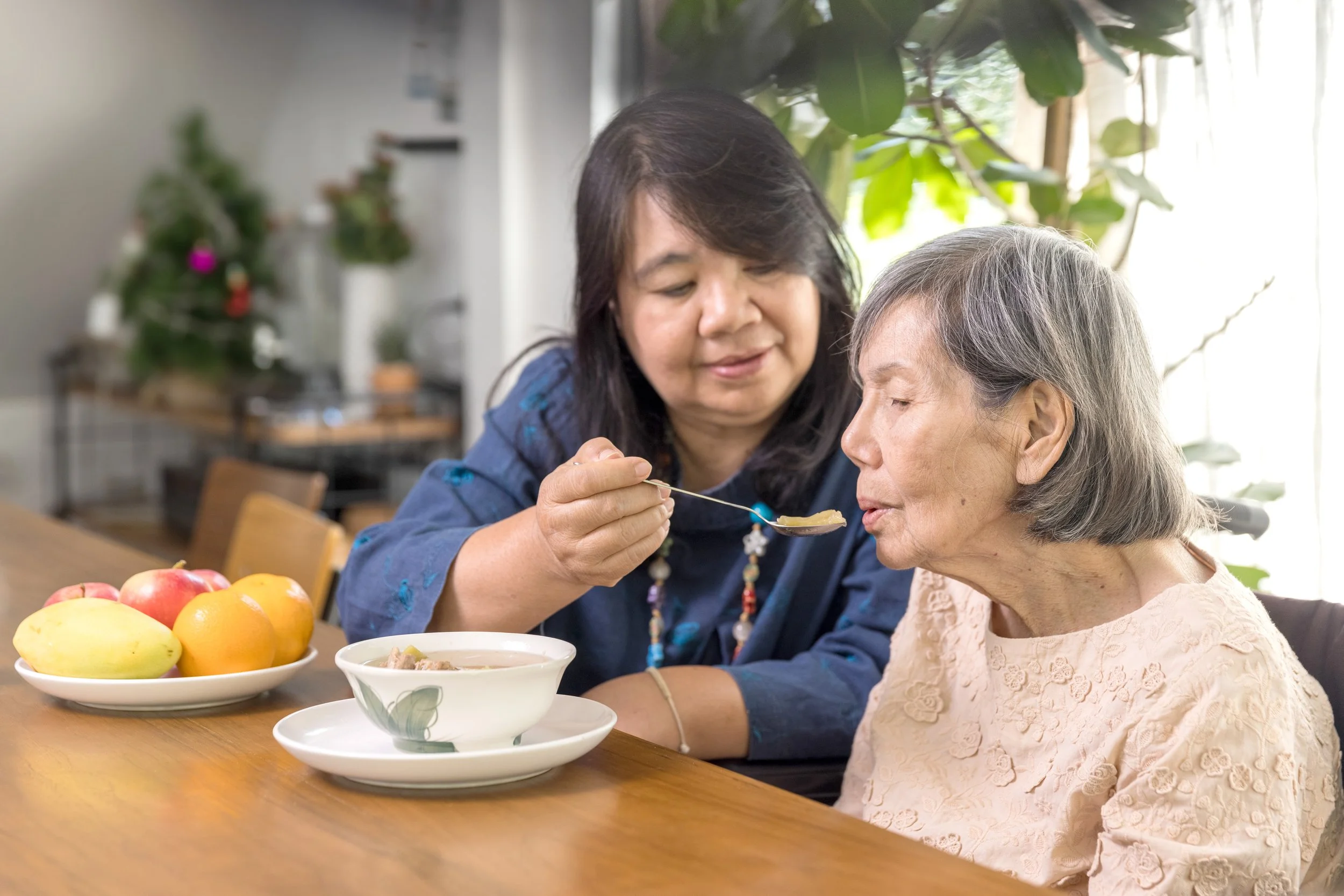 An elderly woman feeding an elderly man in a bright kitchen with fresh vegetables on the counter.