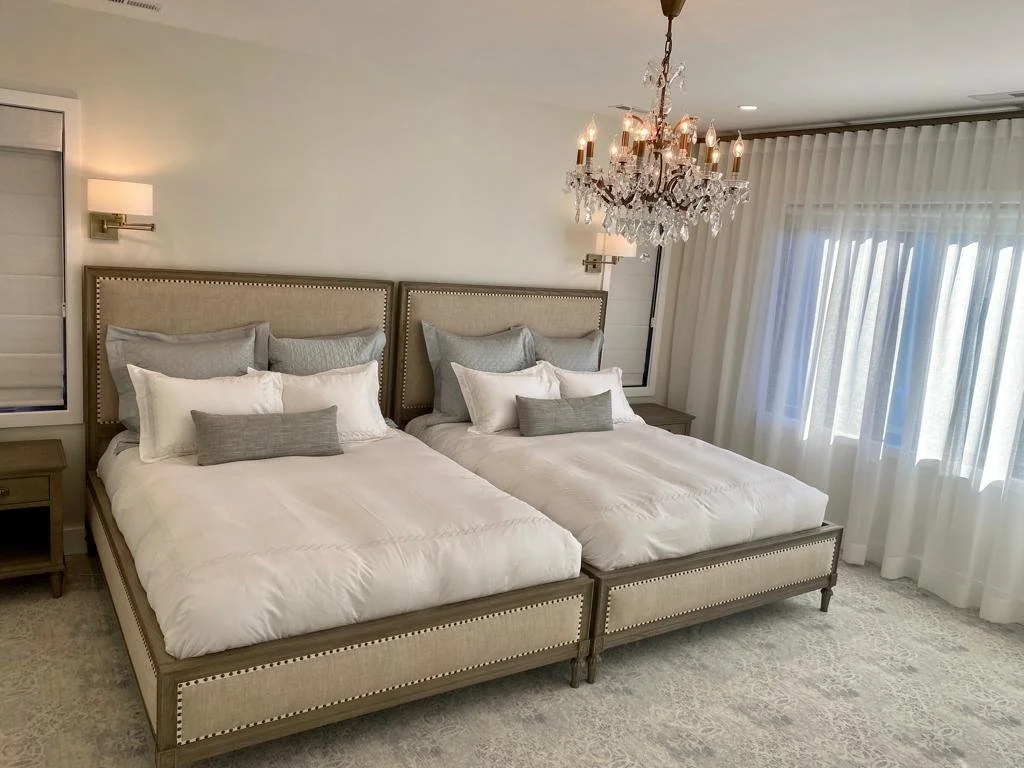 A bedroom with two beds, beige upholstered headboards, white bedding, and various gray and white pillows. There is a chandelier hanging from the ceiling, a window with white curtains, and a small wooden nightstand.