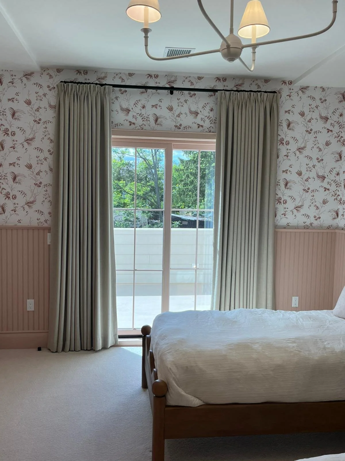 A bedroom with a bed, cream-colored curtains, floral-patterned wallpaper, and a sliding glass door leading to a patio, view of trees outside.