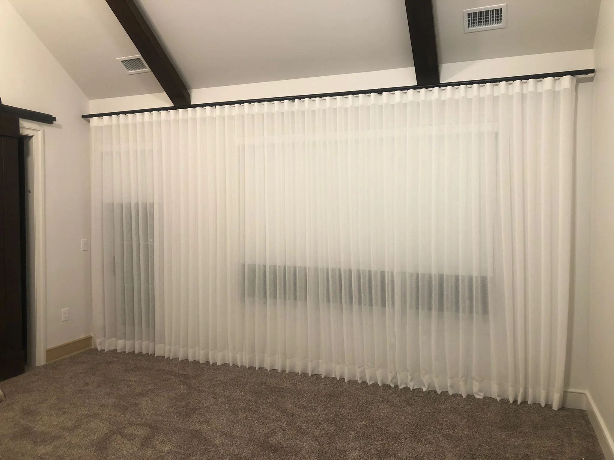 A room with white pleated curtains covering large windows. The room has a gray carpeted floor, white walls, a black ceiling beam, and a black curtain rod. There are vents on the ceiling and wall.