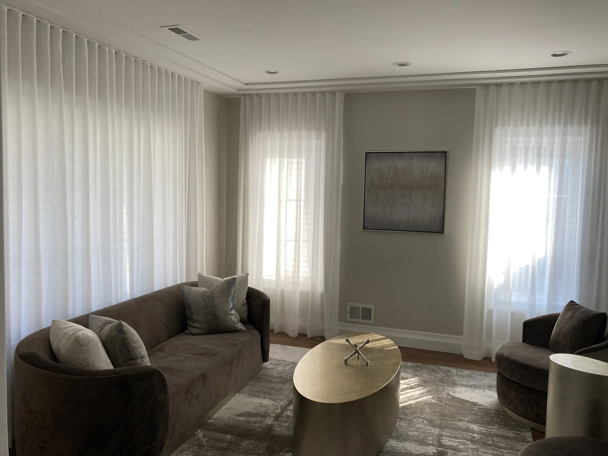 Living room with beige velvet sofa, gray throw pillows, round ottoman coffee table, and armchair, all with light-colored curtains and a wall hanging art piece.
