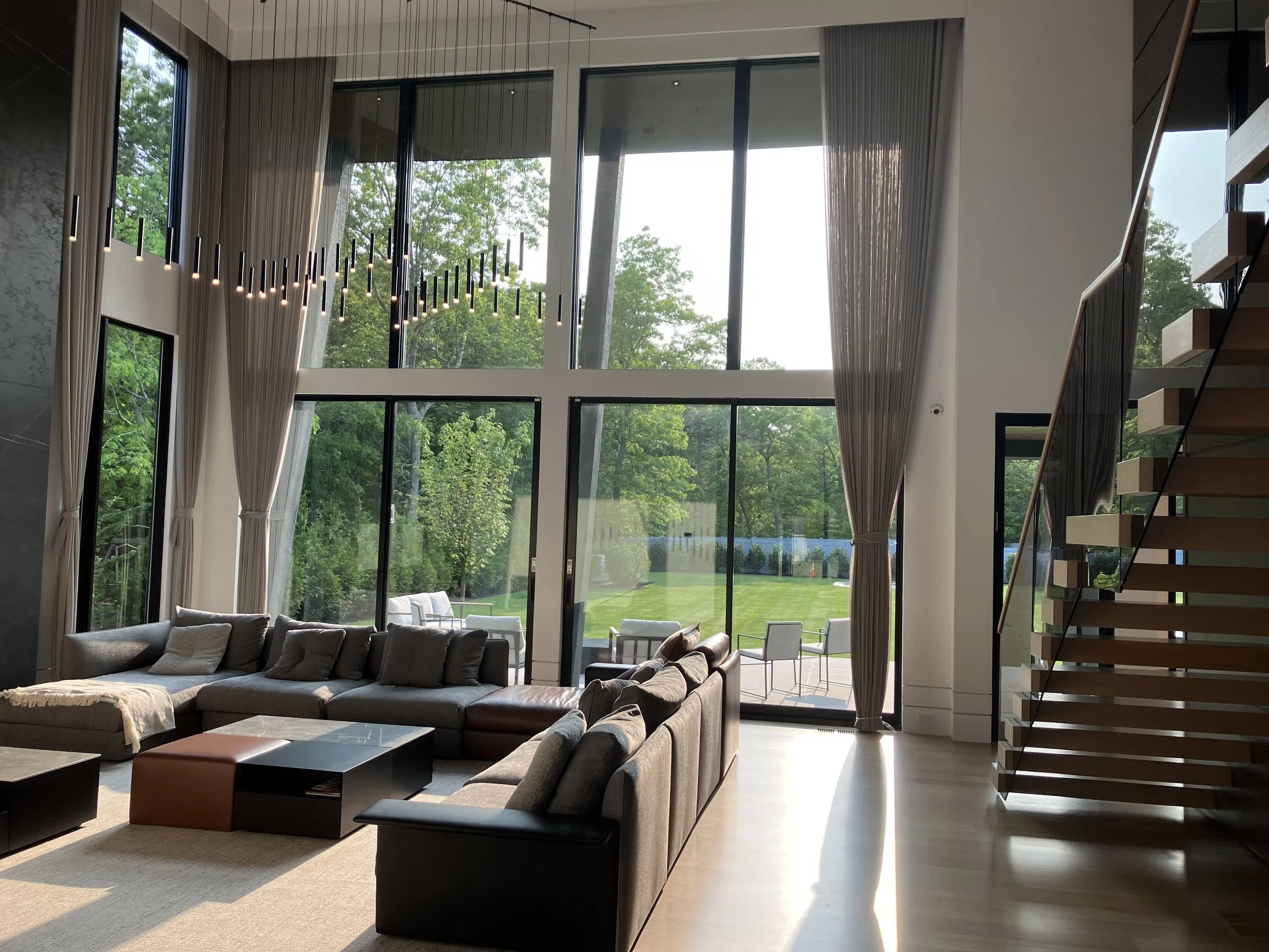Modern living room with large floor-to-ceiling windows, beige curtains, gray sectional sofa with cushions, black and brown coffee tables, and a staircase with wooden steps and a metal railing.