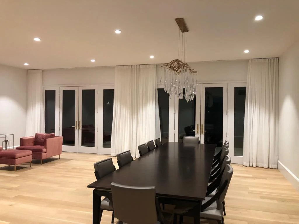 Modern dining room with a long dark wood table, black chairs, white curtains, and a chandelier, with a seating area featuring pink armchairs and a small table.