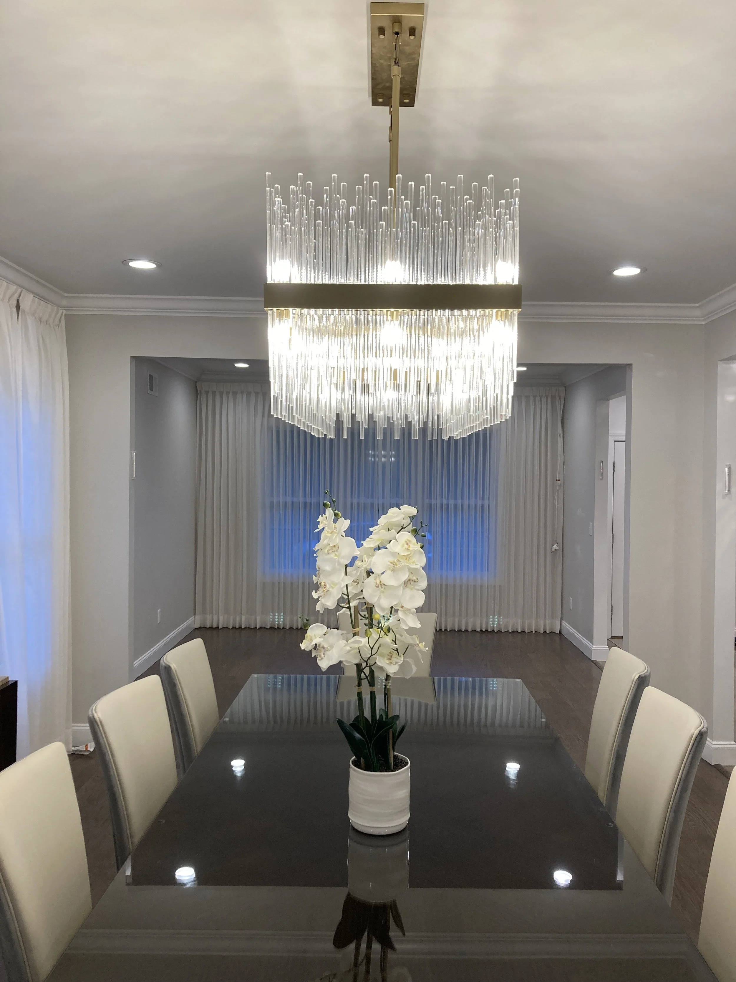 A dining room with a dark wooden table, white chairs, a white potted orchid centerpiece, a large chandelier above, and curtains covering windows in the background.