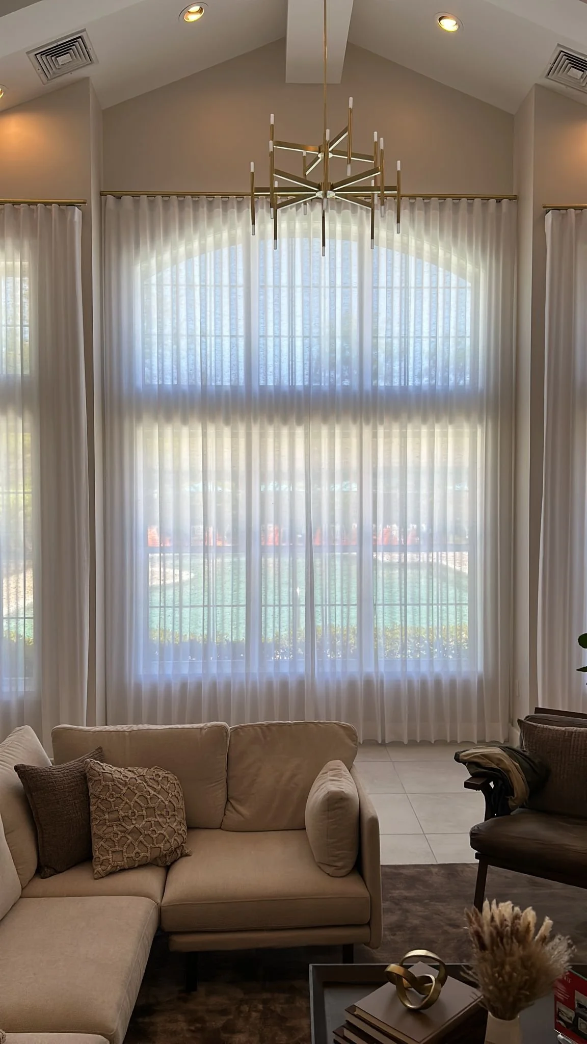 Living room with large arched window covered with sheer curtains, a beige sectional sofa with decorative pillows, a chandelier, and a coffee table with decorative items.