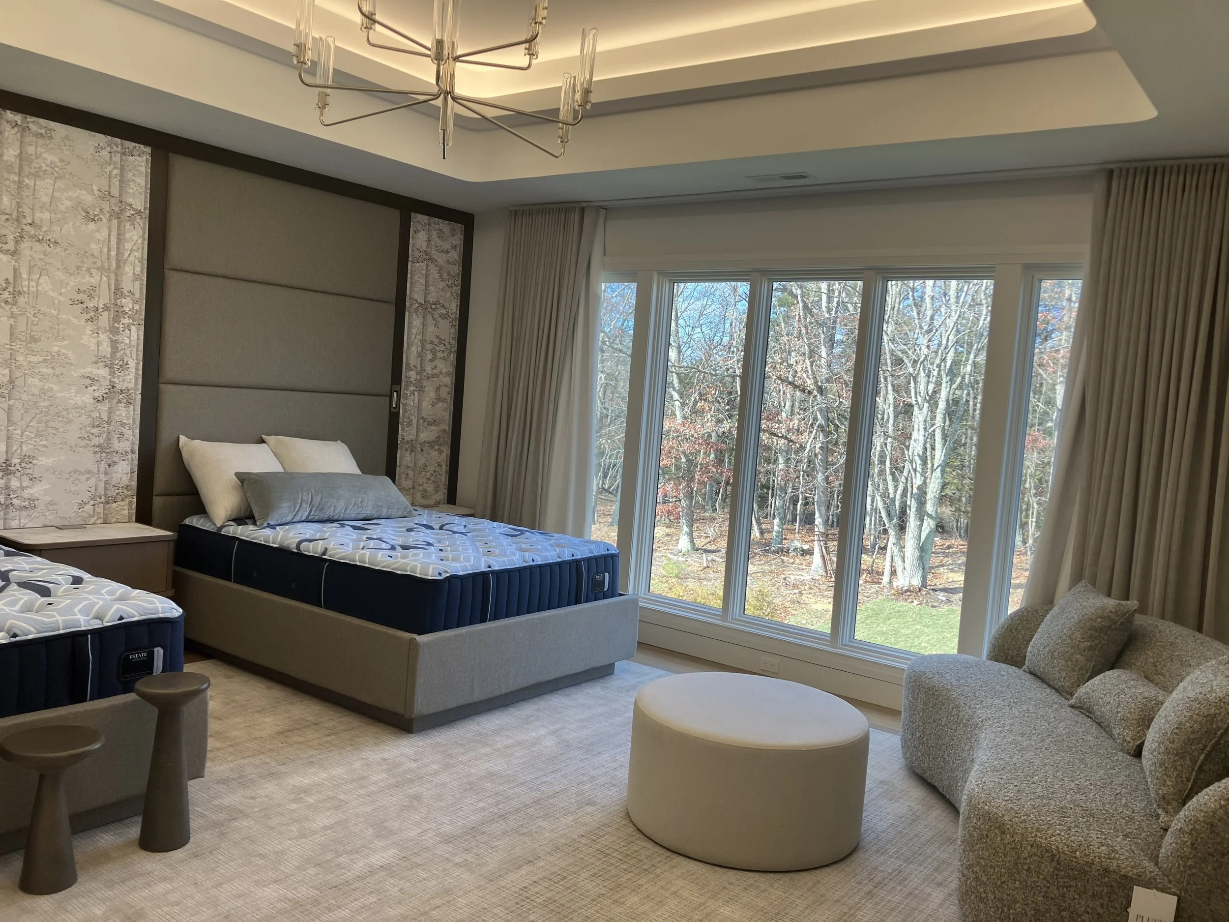 A modern bedroom with a large window overlooking a wooded outdoor area, featuring a bed with a gray upholstered headboard, a side table, a gray armchair with pillows, a white ottoman, and two small stools.