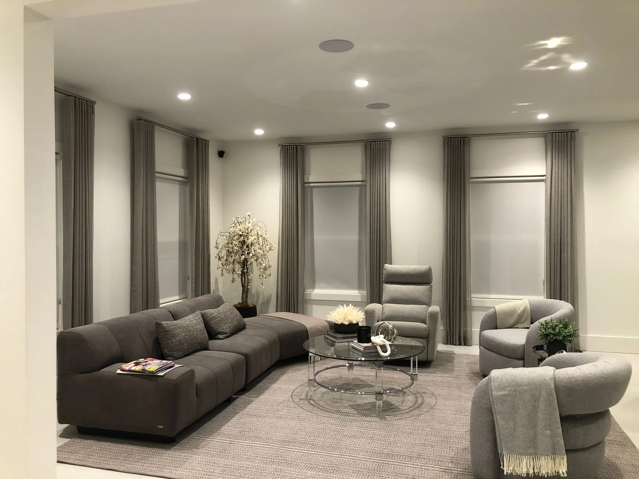 Modern living room with gray upholstered sofa, matching armchairs, a glass coffee table, and tall windows with gray curtains.