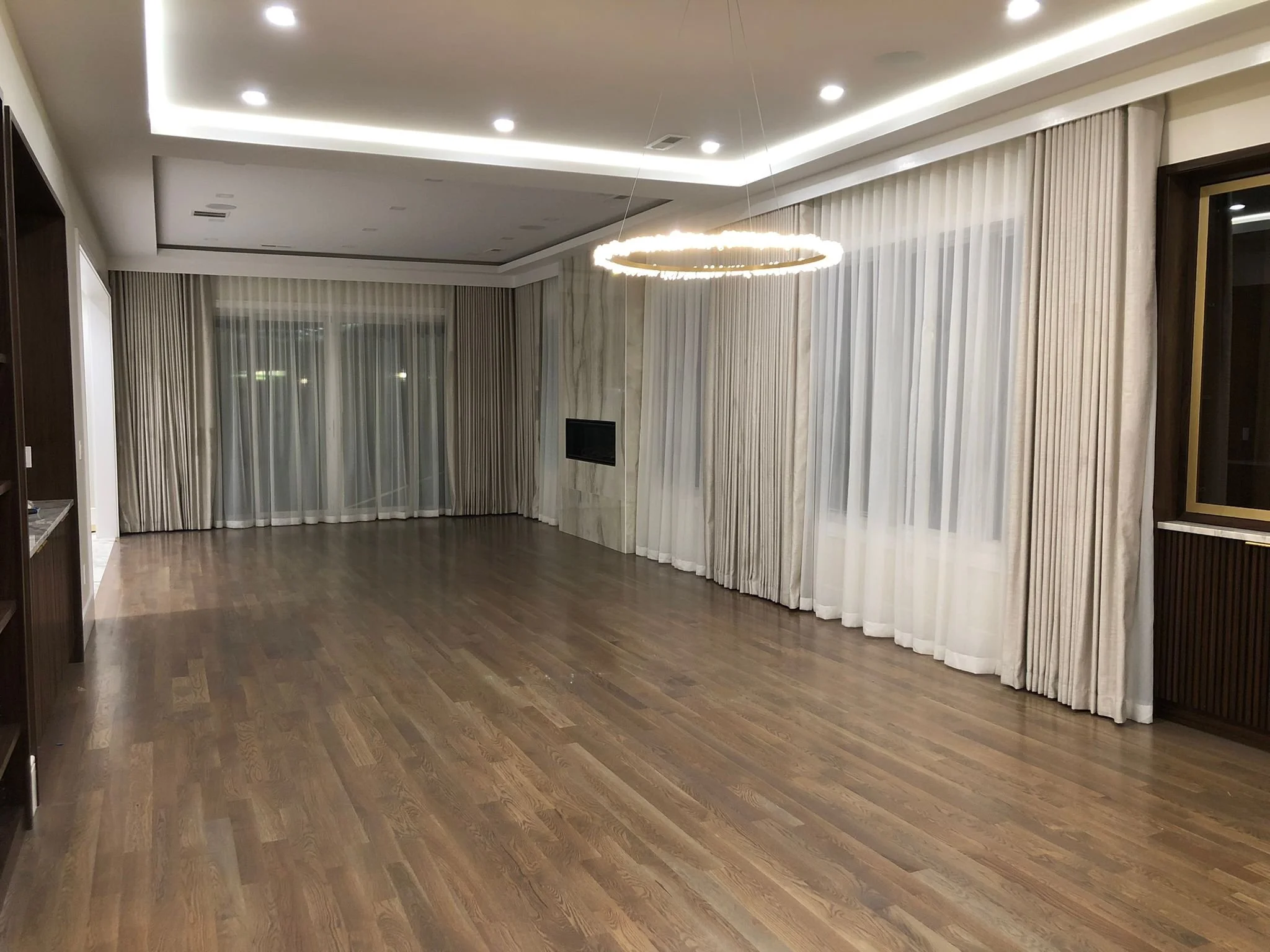 Empty living room with hardwood floors, large windows with white curtains, and a ceiling light fixture.